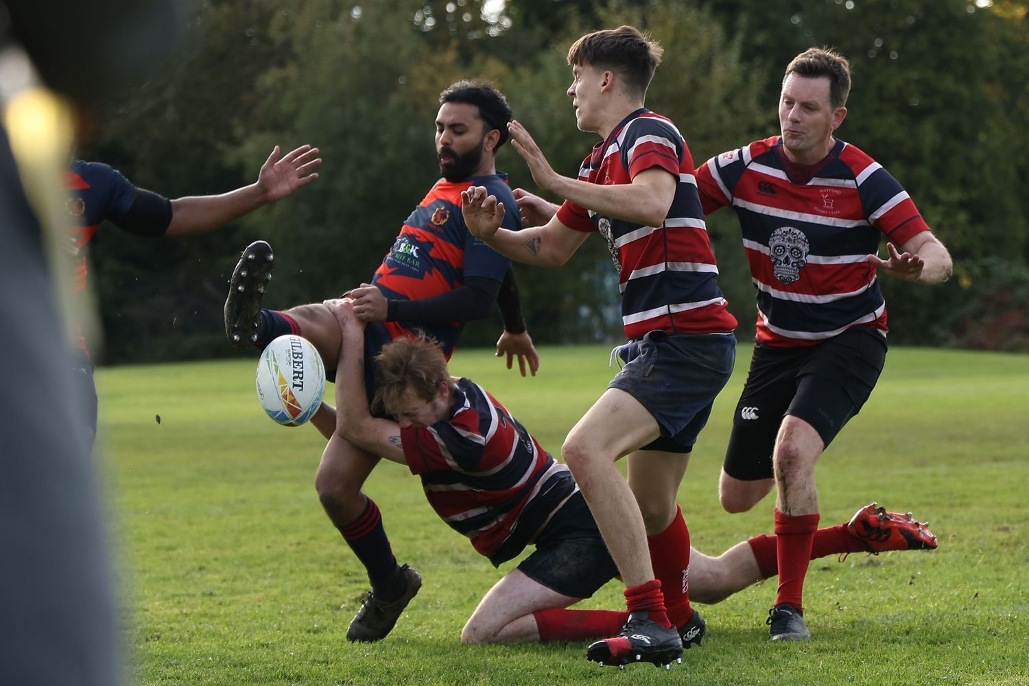 Rugby players engage in a dramatic dive tackle during an outdoor match as teammates watch in the background.