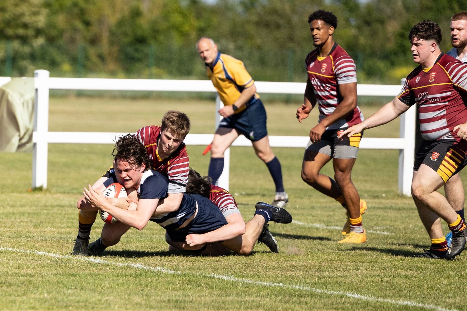 Rugby players compete for the ball during an intense tackle on a sunny grass field with white boundary posts visible.