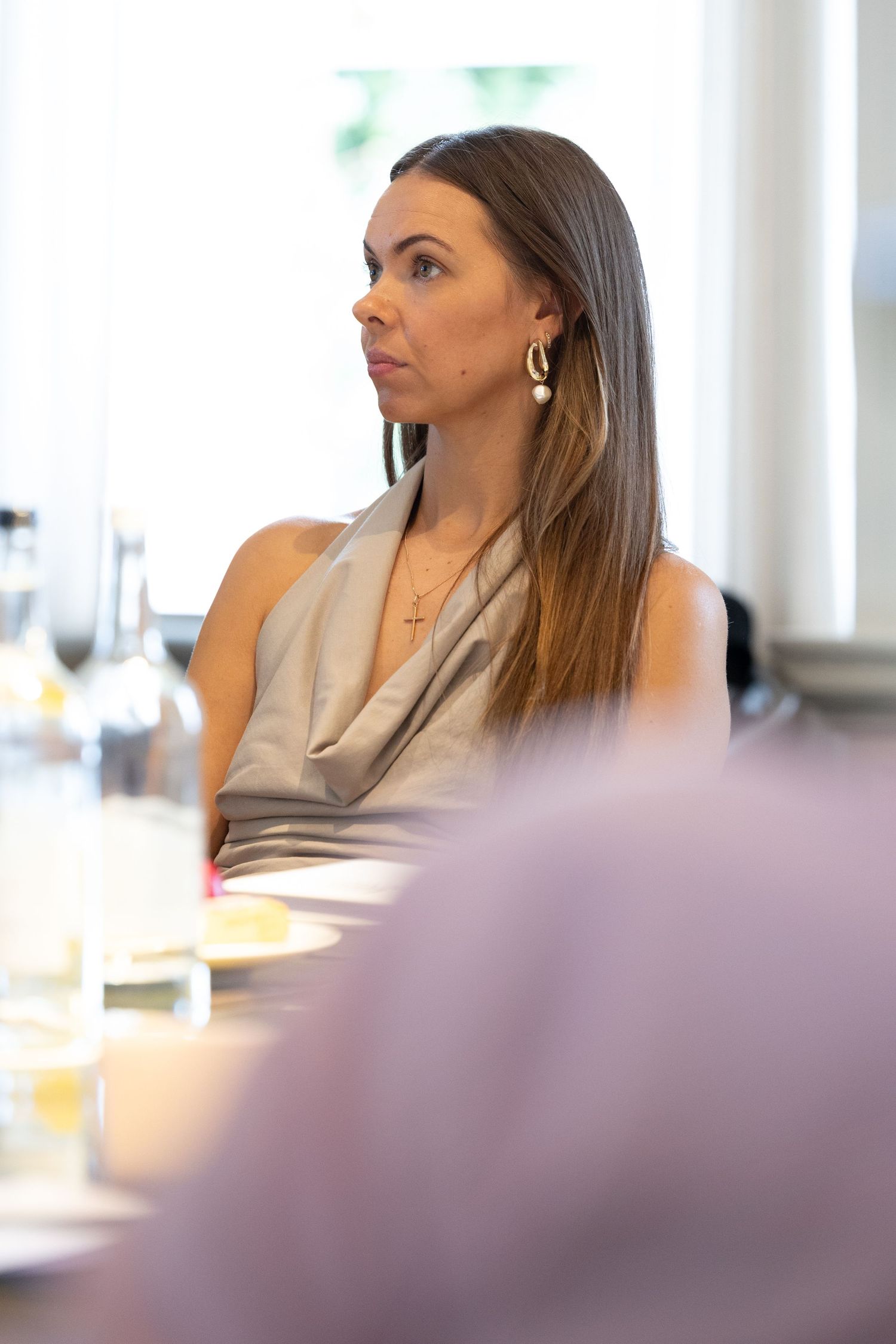 Woman in beige sleeveless top and hoop earrings sits at dining table looking to the side.