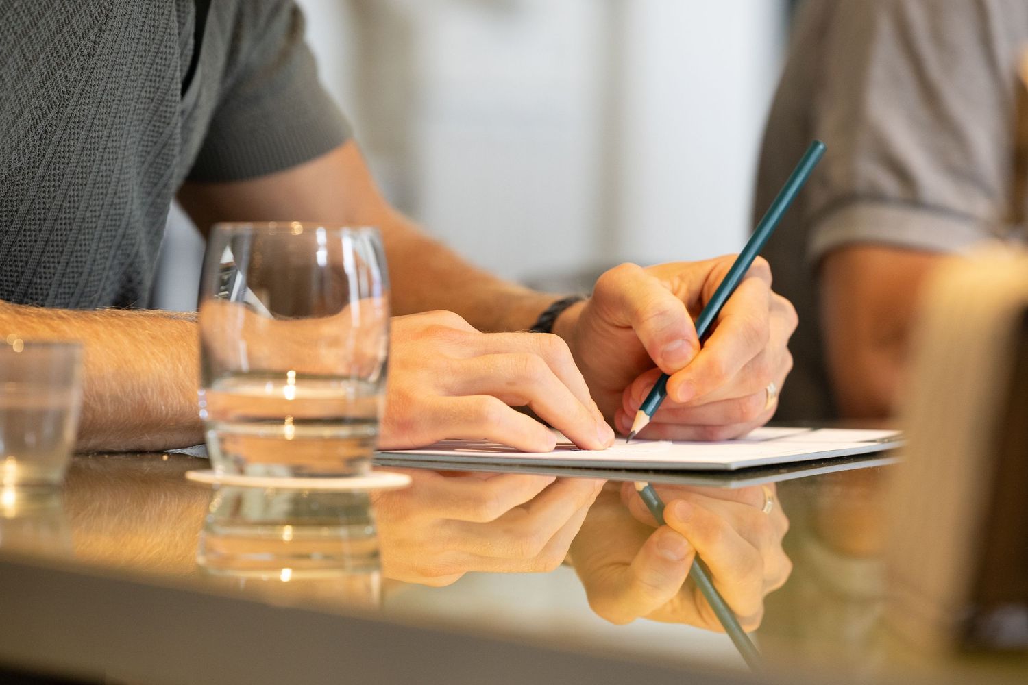 Person writing notes on paper with glass of water on reflective table surface in meeting room setting.