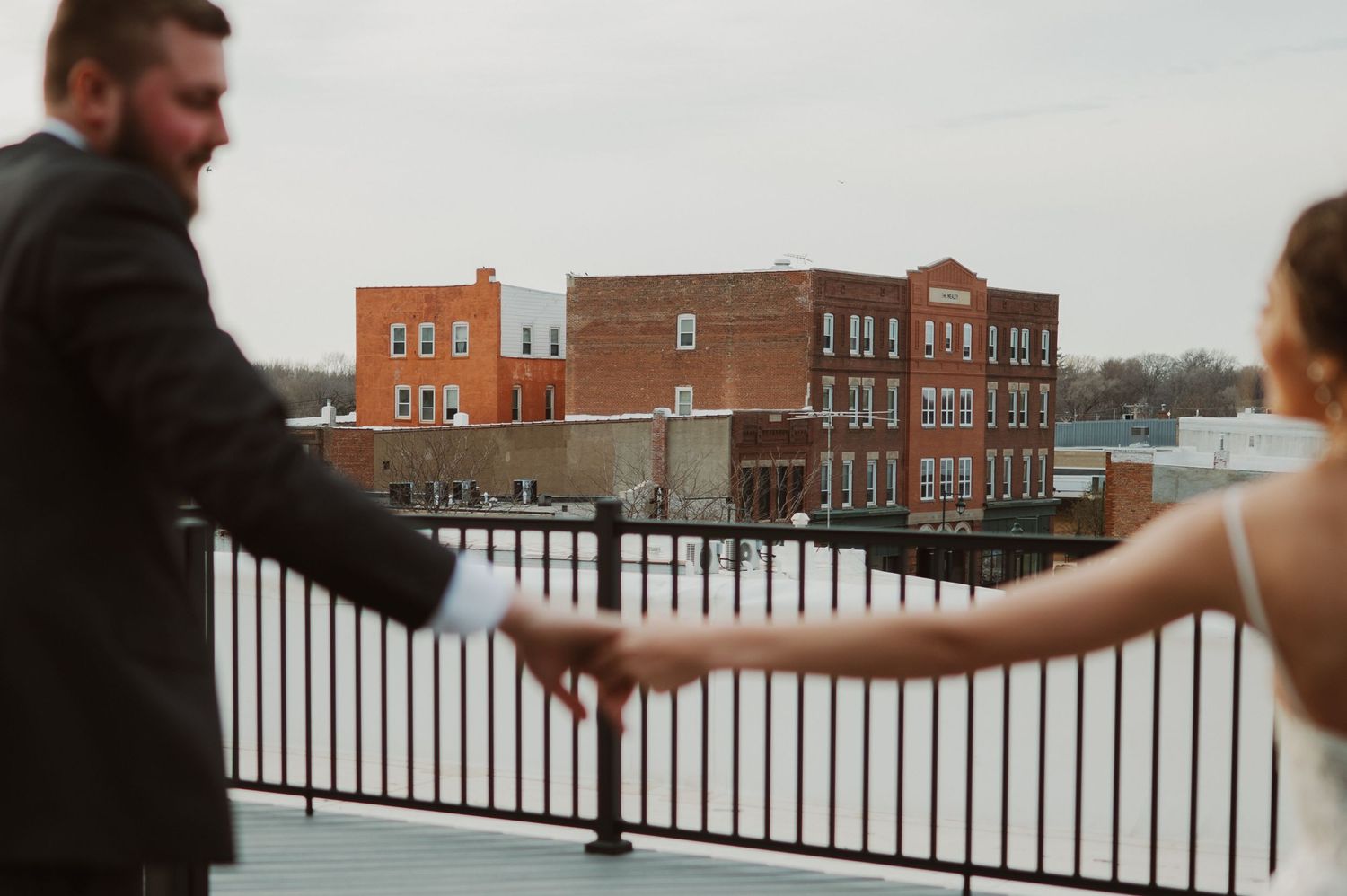 Two people hold hands while standing on a snowy rooftop balcony with a brick apartment building in the background.