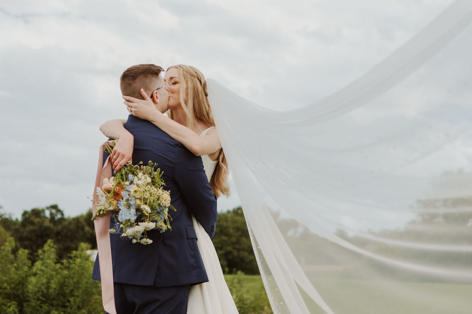 A couple embraces during their outdoor wedding ceremony with a flowing white veil in the breeze.