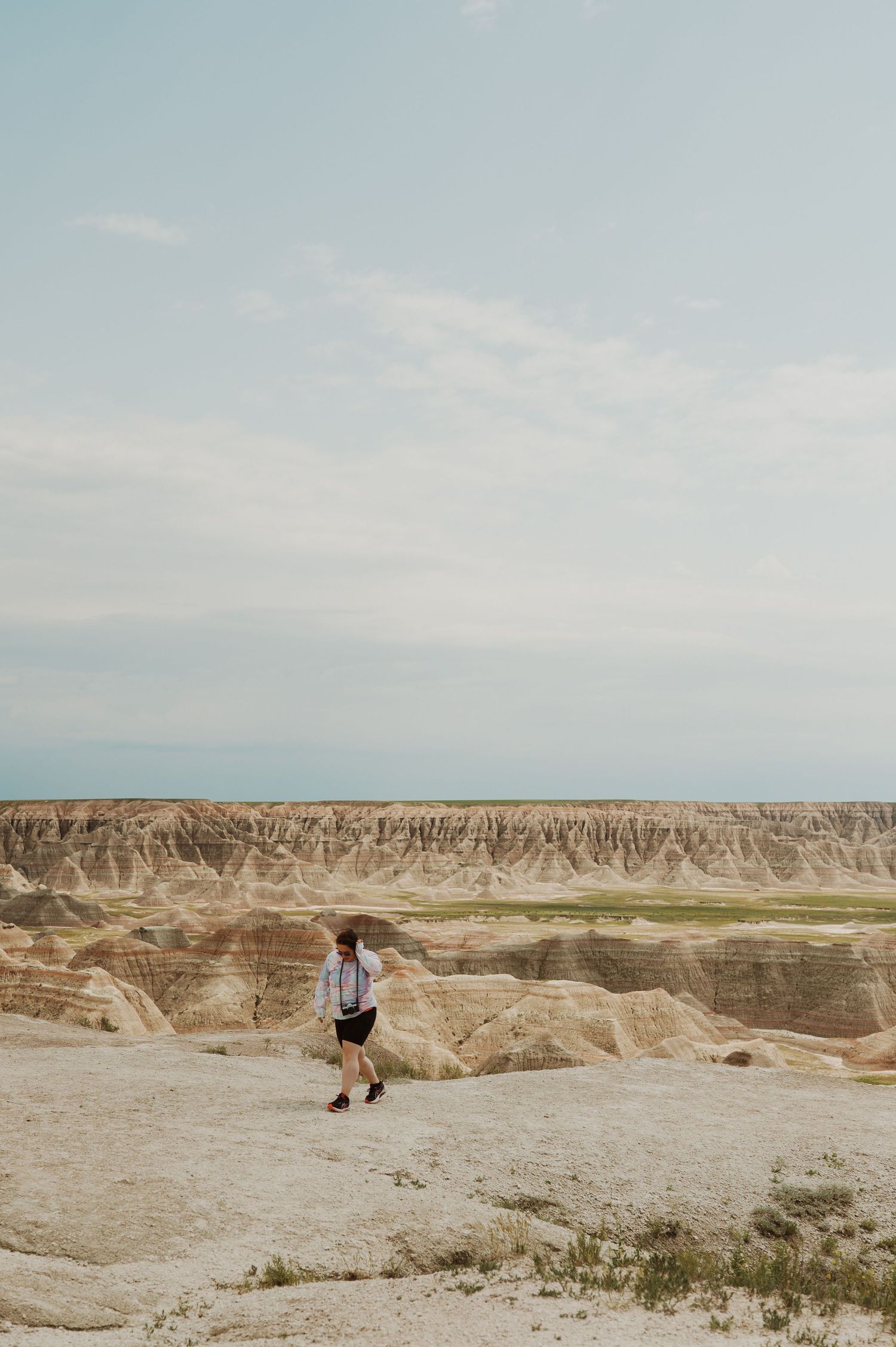 A lone hiker walks along rocky terrain in the Badlands National Park under a vast cloudy sky.