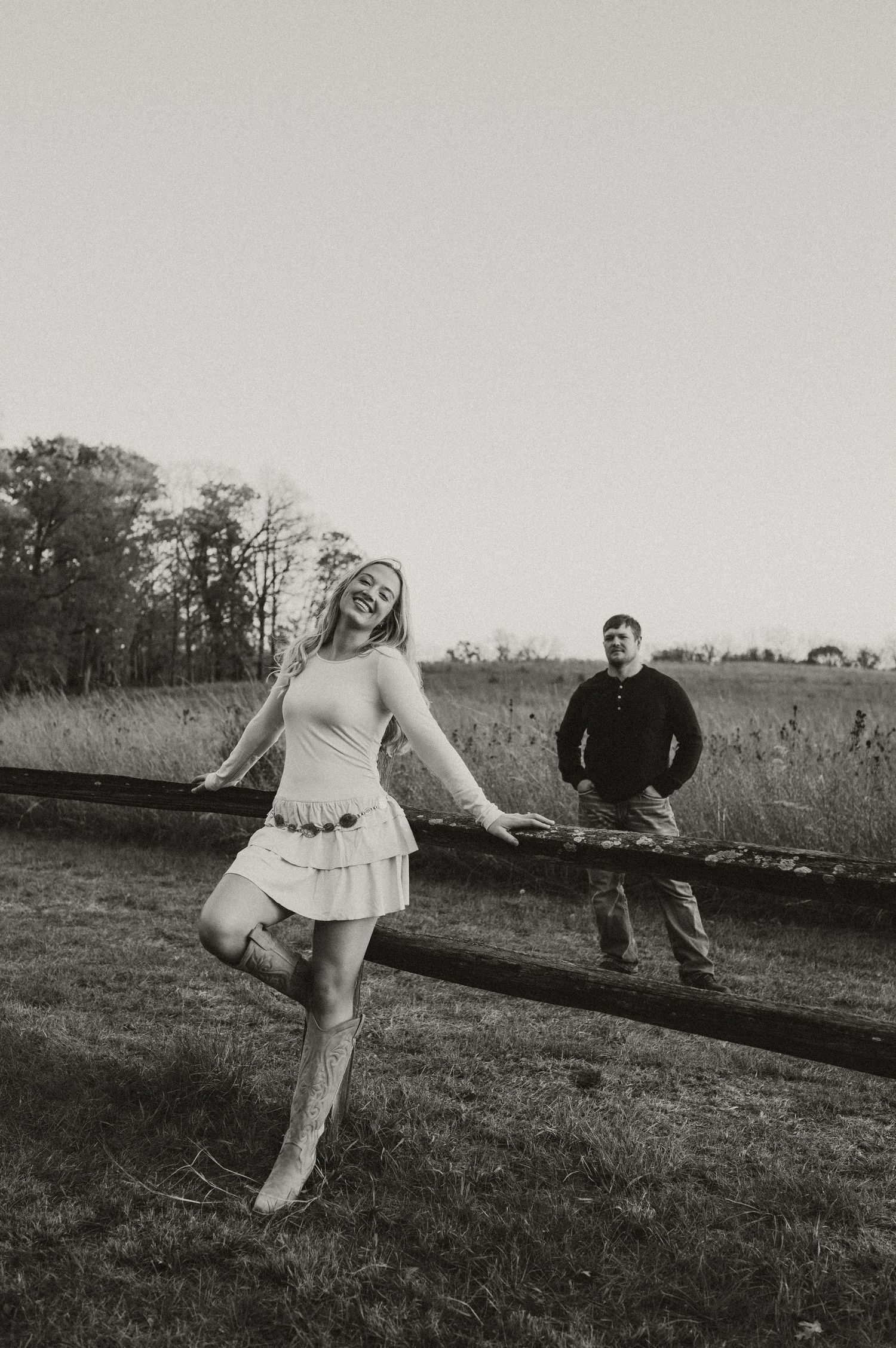 Black and white photo of a couple posing by a wooden fence in a rural field setting.