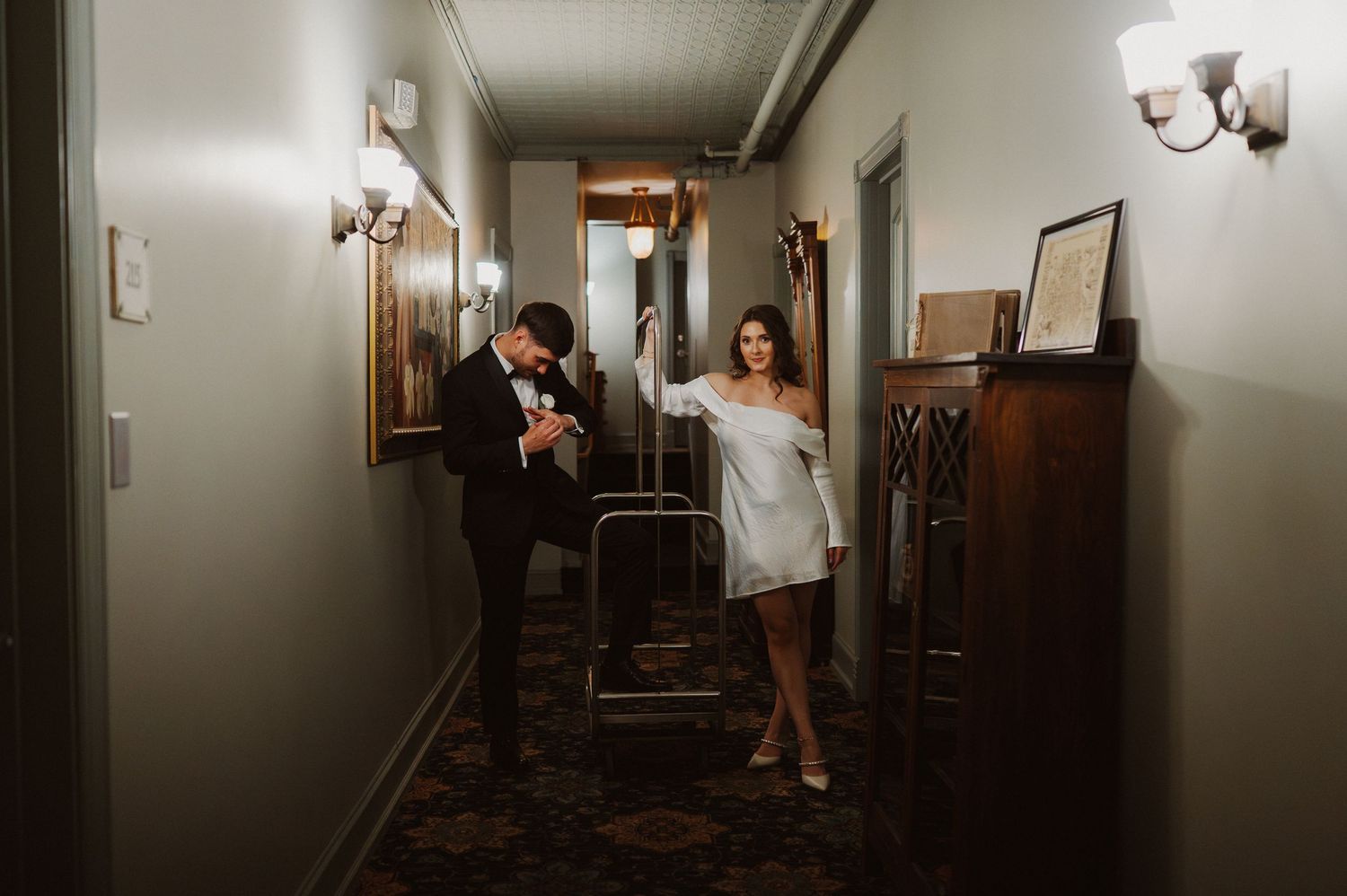 A couple in wedding attire playfully poses in a dimly lit vintage hotel hallway with antique furniture.