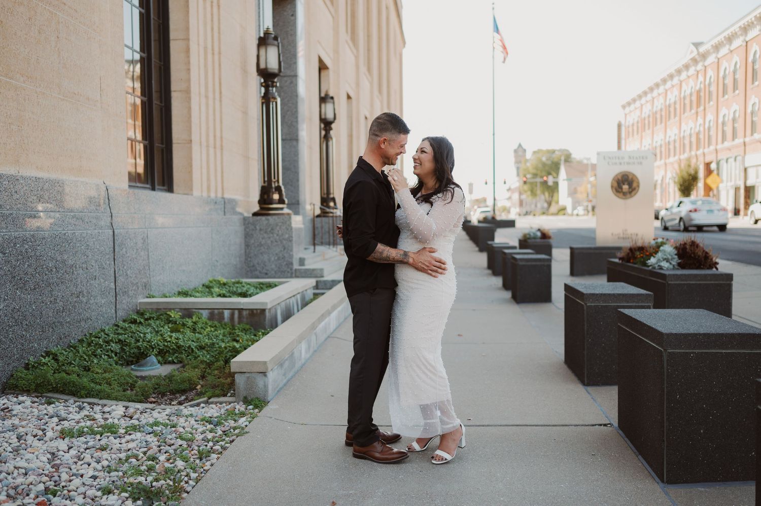 A couple in formal attire embrace on a city sidewalk next to modern outdoor seating and architectural details.
