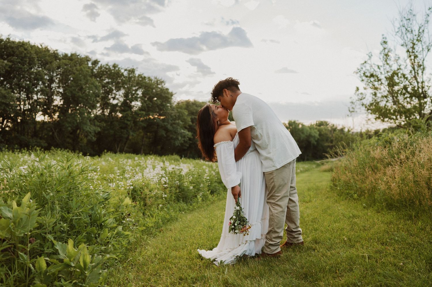 A romantic couple embraces in a lush green meadow on a cloudy summer day.