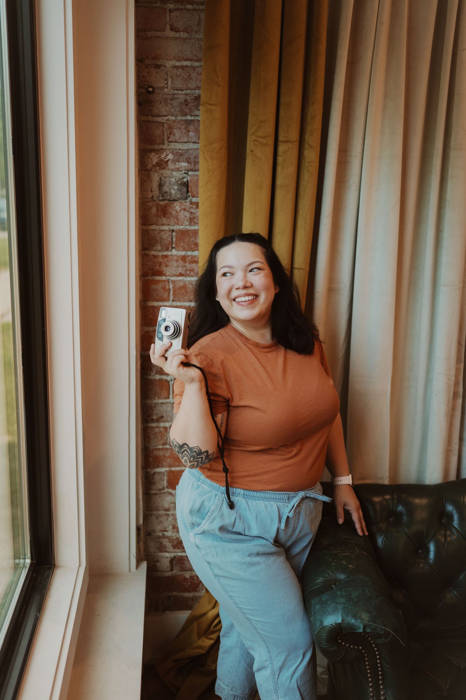 Person in rust colored top poses casually against brick wall and curtains.