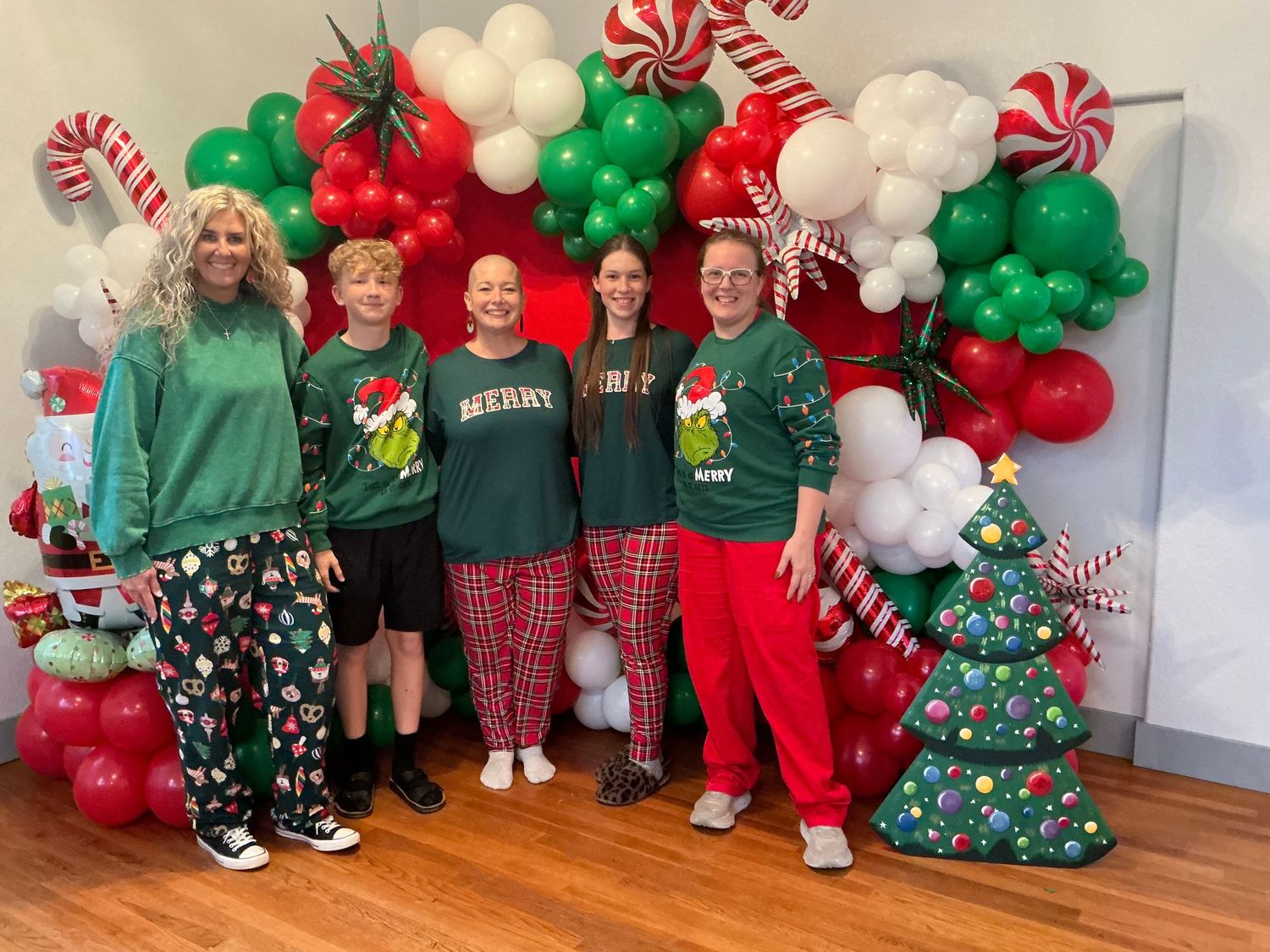 Group of five people in festive Christmas outfits posing in front of a colorful holiday balloon arch display.