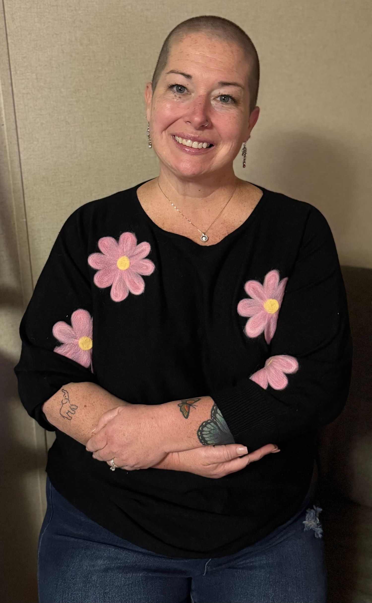Smiling woman with shaved head wearing black floral sweatshirt with pink daisies, arms crossed, showing tattoos.