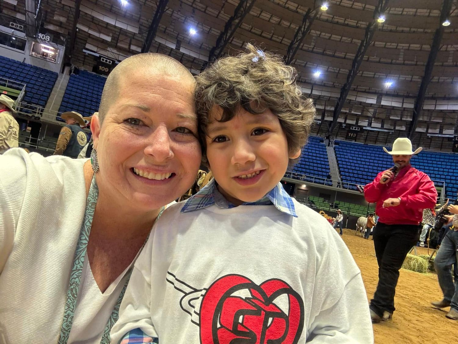 Two people pose for a selfie at an indoor sports arena with stadium seating visible in the background.