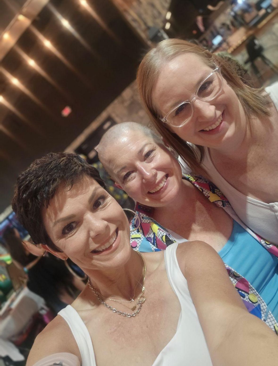 Three smiling women pose together for a selfie indoors at a casual social gathering.