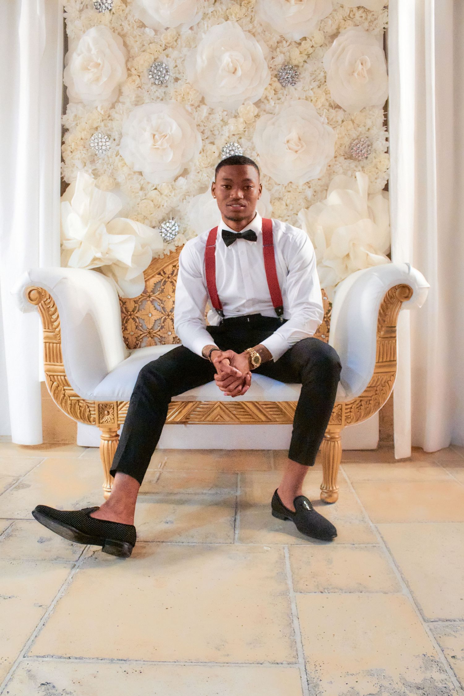 Person in formal attire with suspenders sitting on an elegant white chaise lounge.