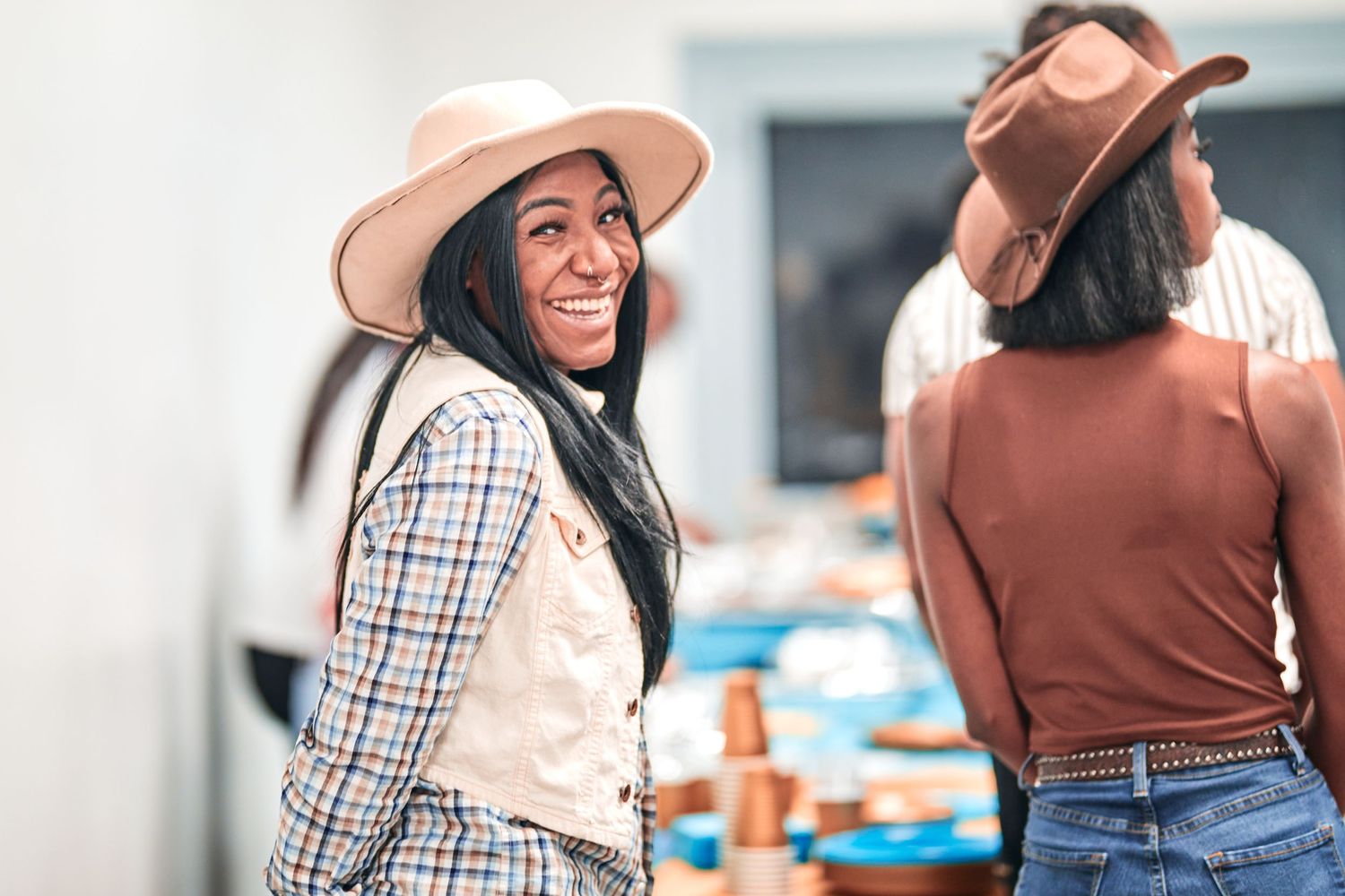 Two people in casual western attire with hats stand in a bright indoor setting.