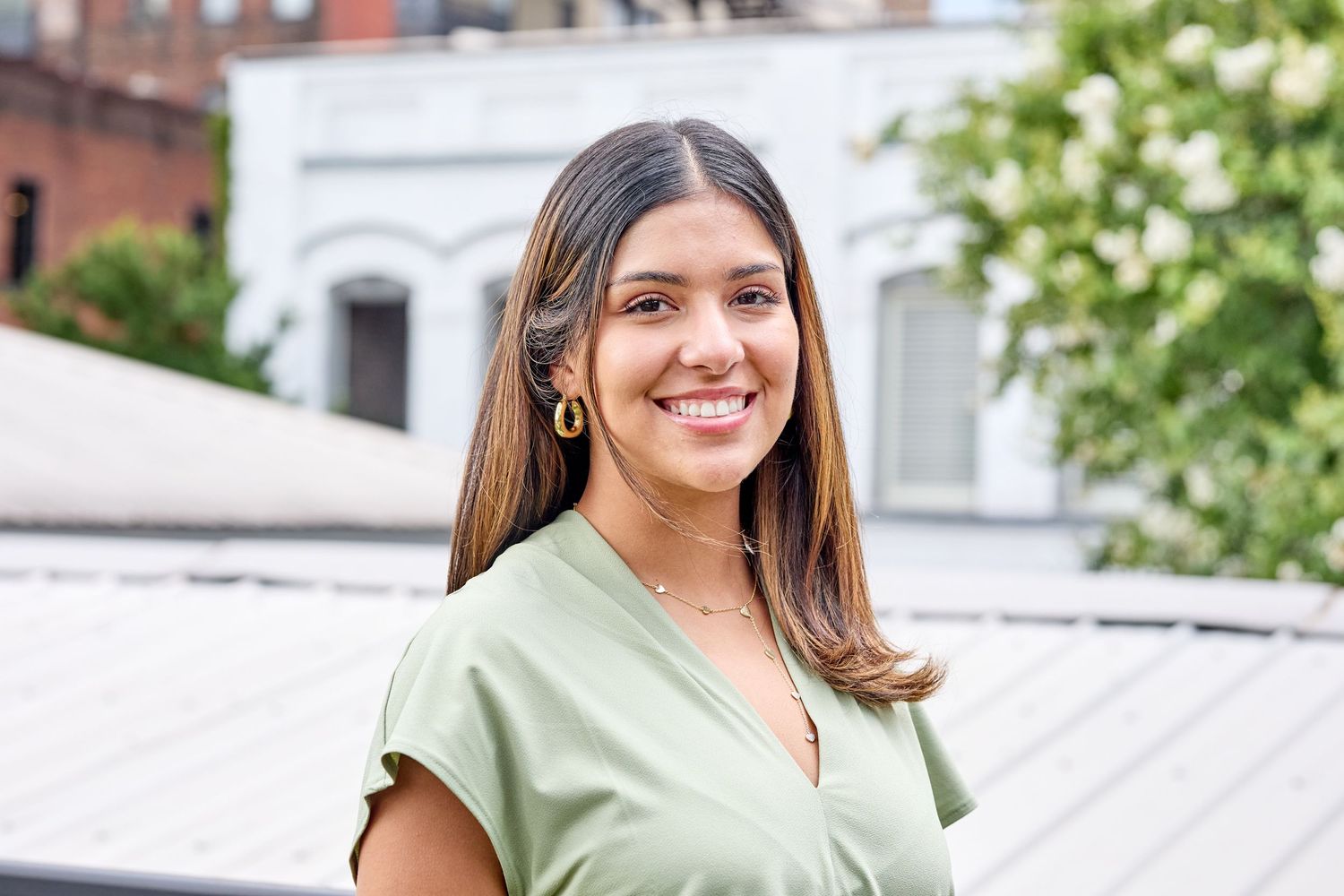 Person in green v-neck top smiling outdoors with urban architecture in background.