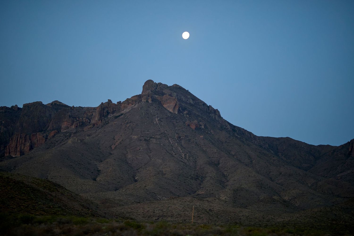Full moon rising over rugged mountain peak during dusk against clear blue sky.