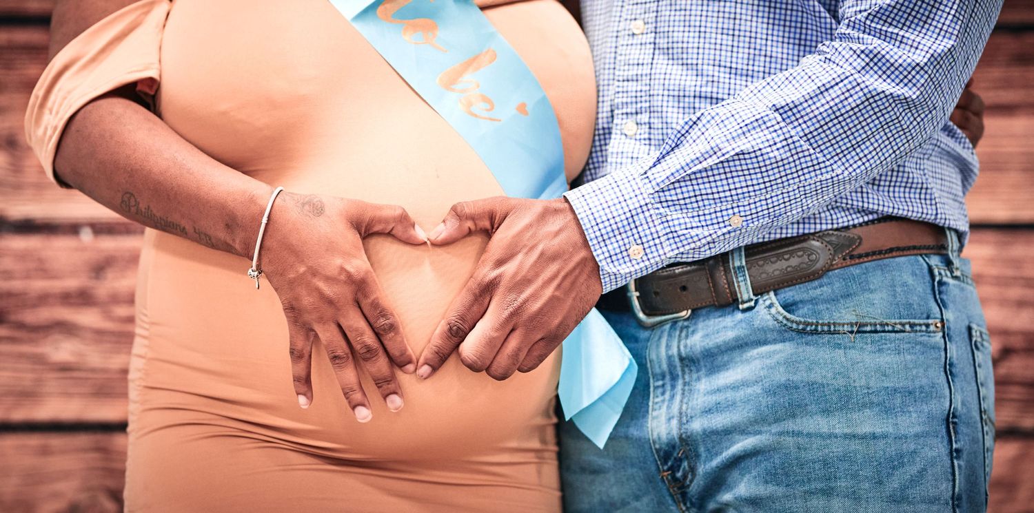 A close-up of hands forming a heart shape over a pregnant belly.