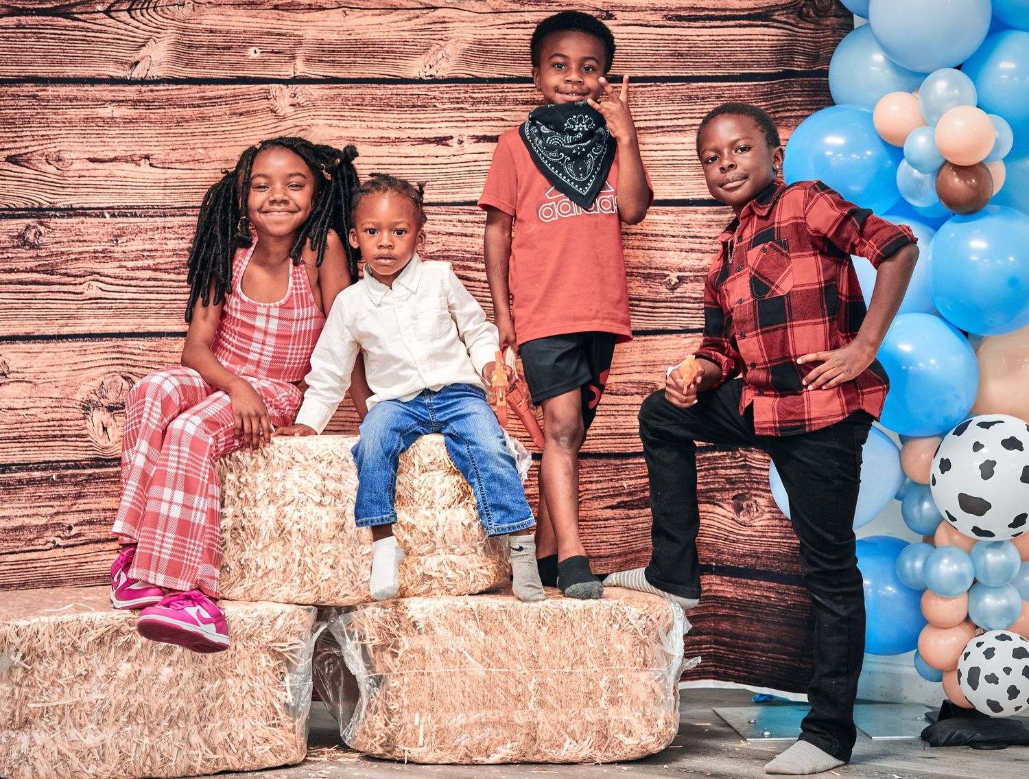 Children standing on hay bales with blue and peach balloons against rustic wooden background.