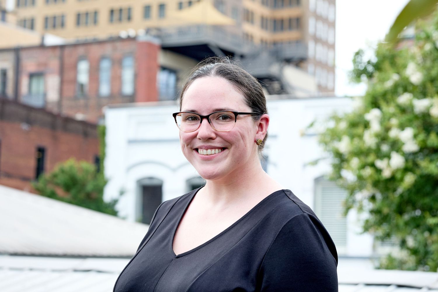 Person in black top wearing glasses smiling outdoors with buildings in background.