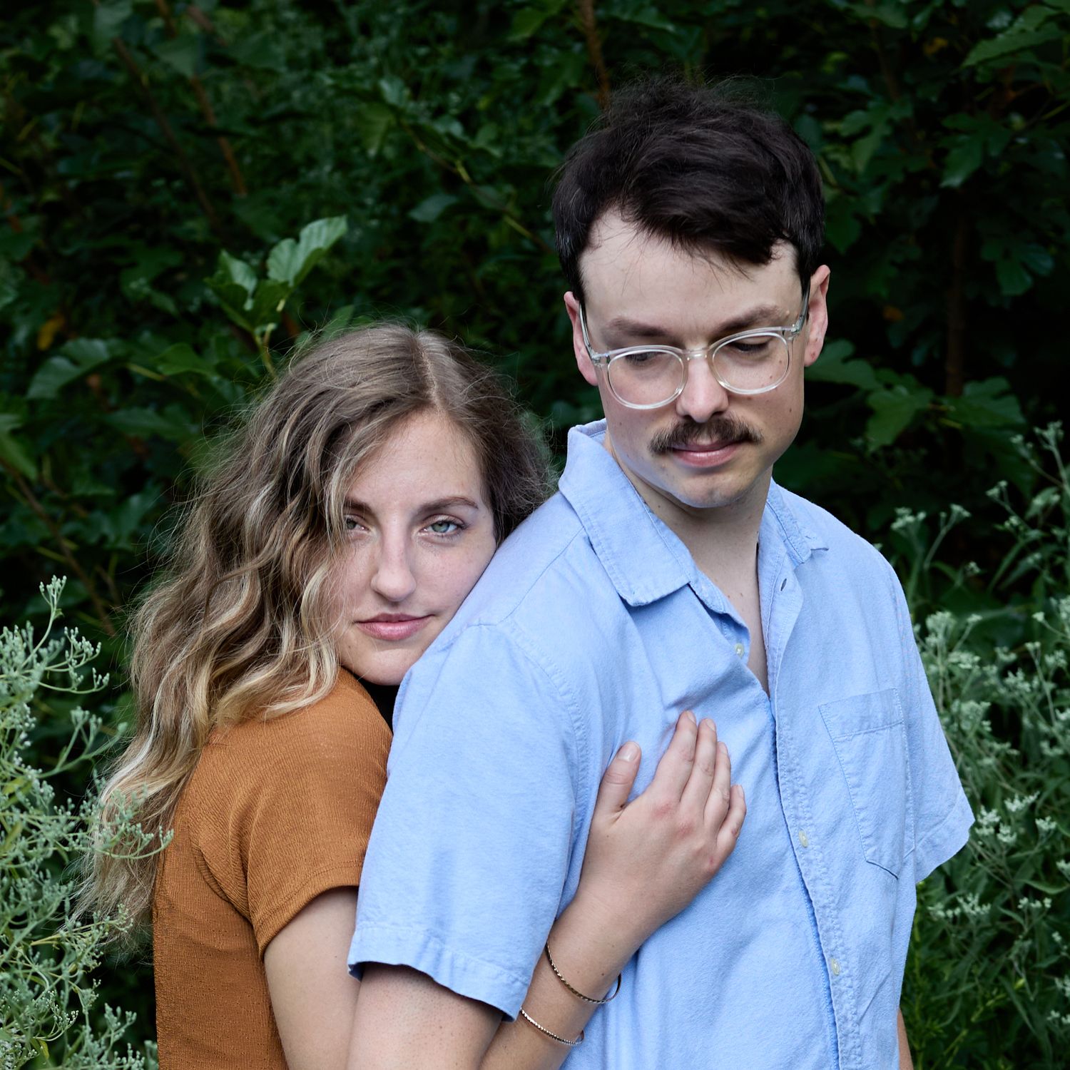 A couple in casual attire standing together against green foliage background.
