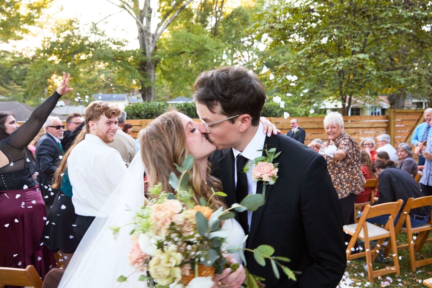 A bride and groom share a romantic kiss during their outdoor autumn wedding ceremony surrounded by guests.