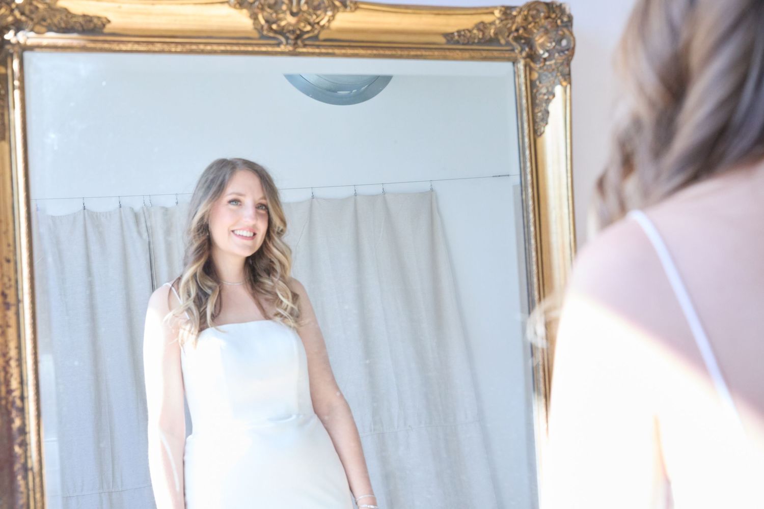 A white wedding dress is reflected in an ornate gold mirror with soft natural lighting.