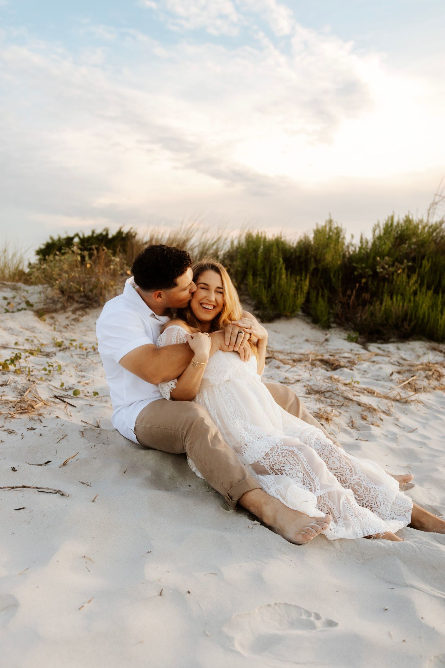 A couple in white and beige clothing embracing on sandy dunes at sunset.