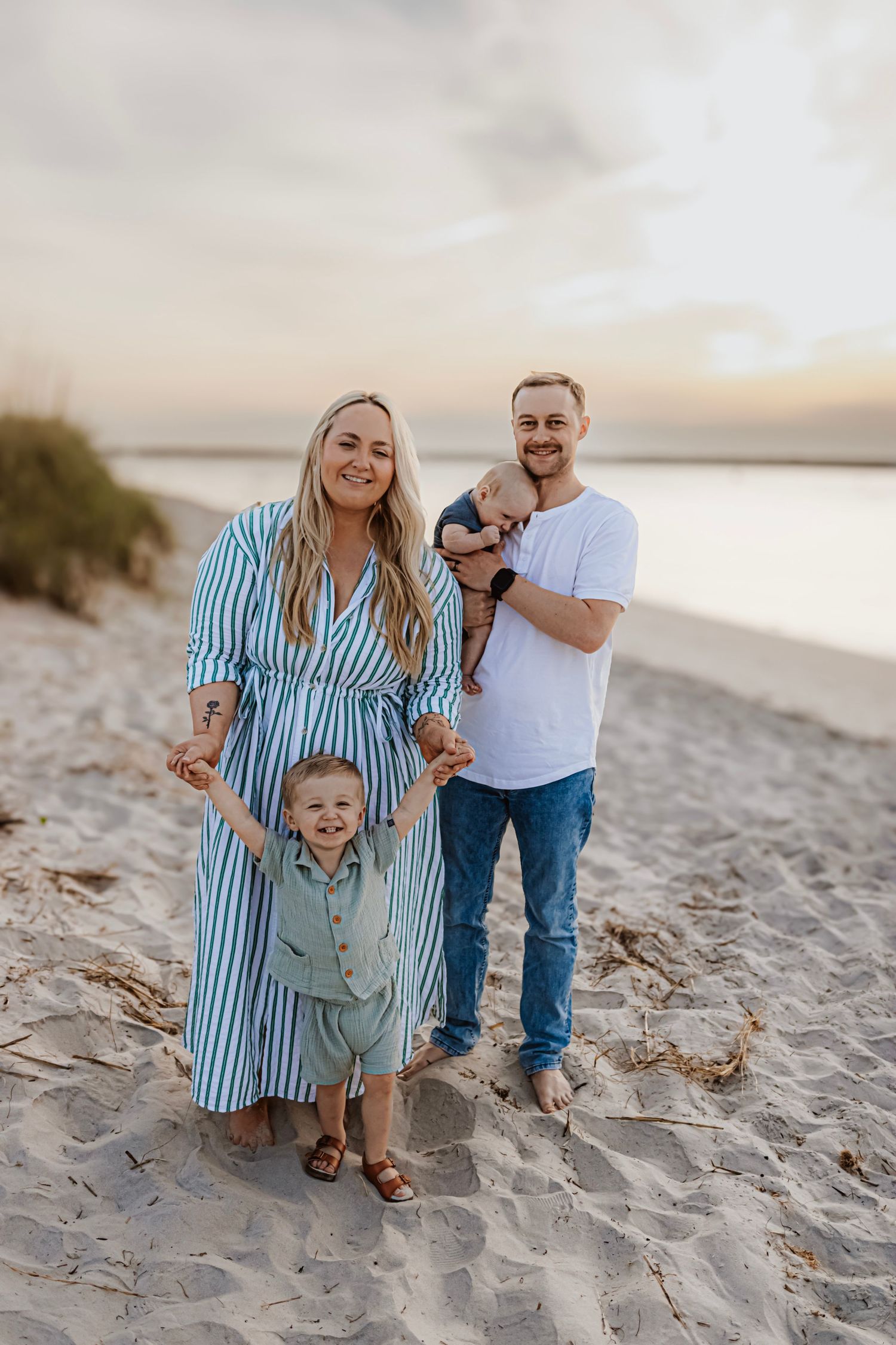 Family of four in coordinating blue and white outfits pose together on sandy beach at sunset.