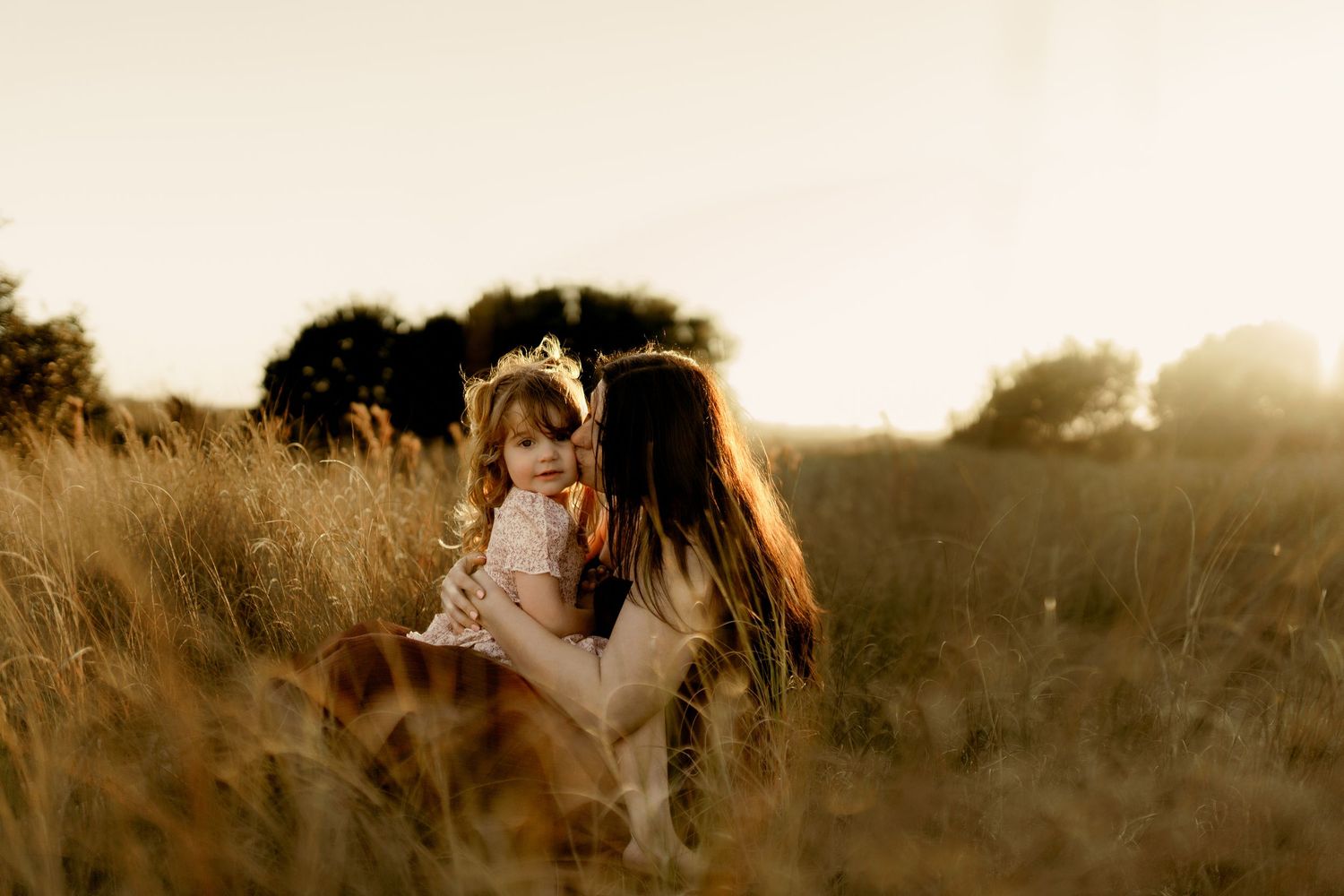 A mother holds her smiling toddler daughter in a golden sunlit field, both wearing white dresses at sunset.
