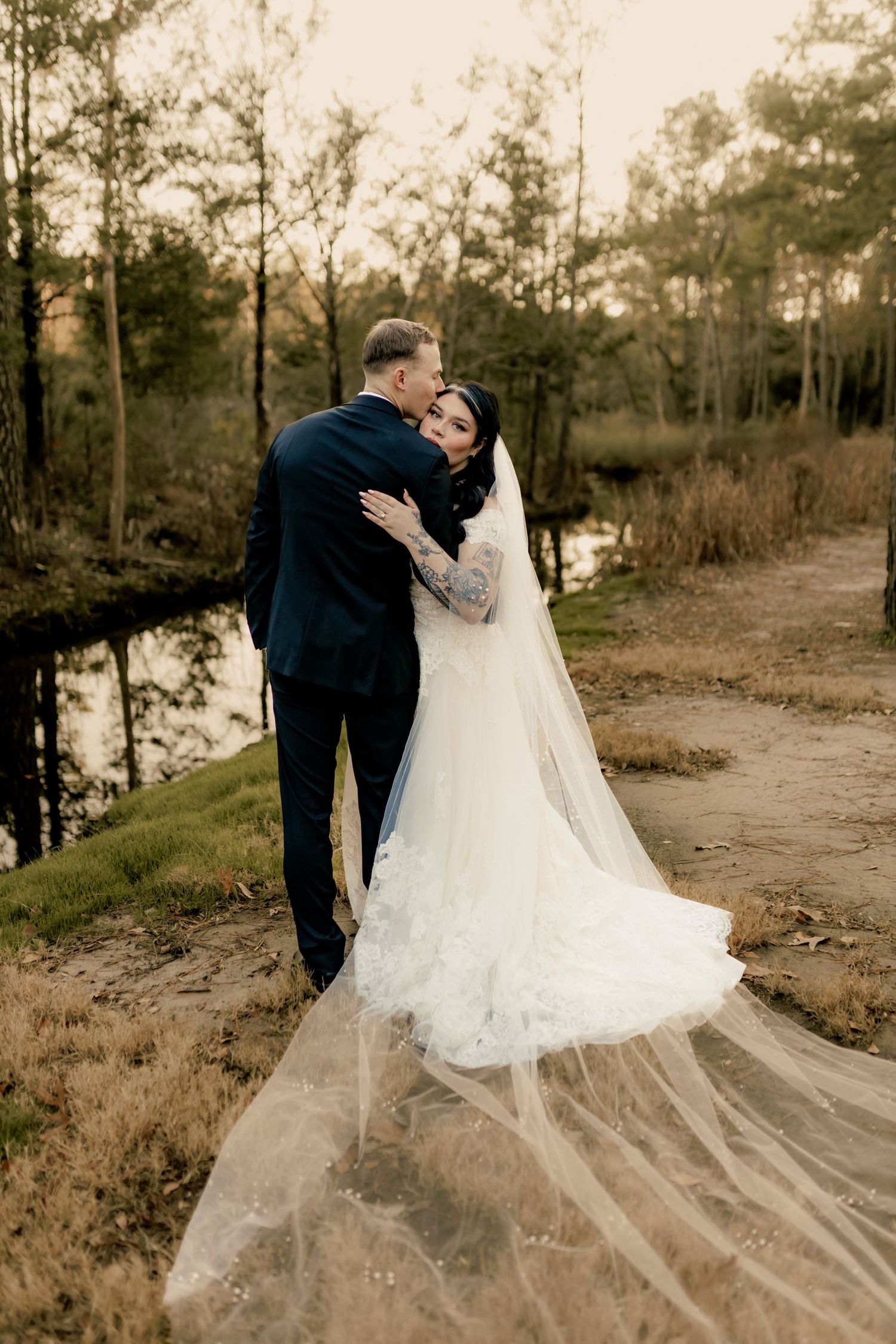 Bride in flowing white gown and veil embraces groom in navy suit near a serene woodland pond at sunset.
