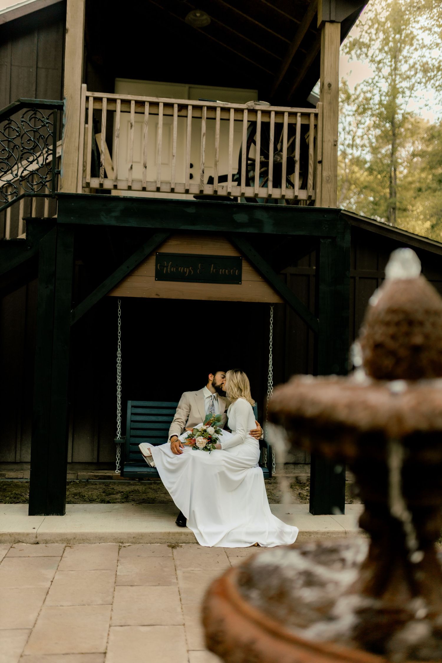 Bride and groom kissing on a swing outside a rustic cabin venue with a stone fountain in the foreground.