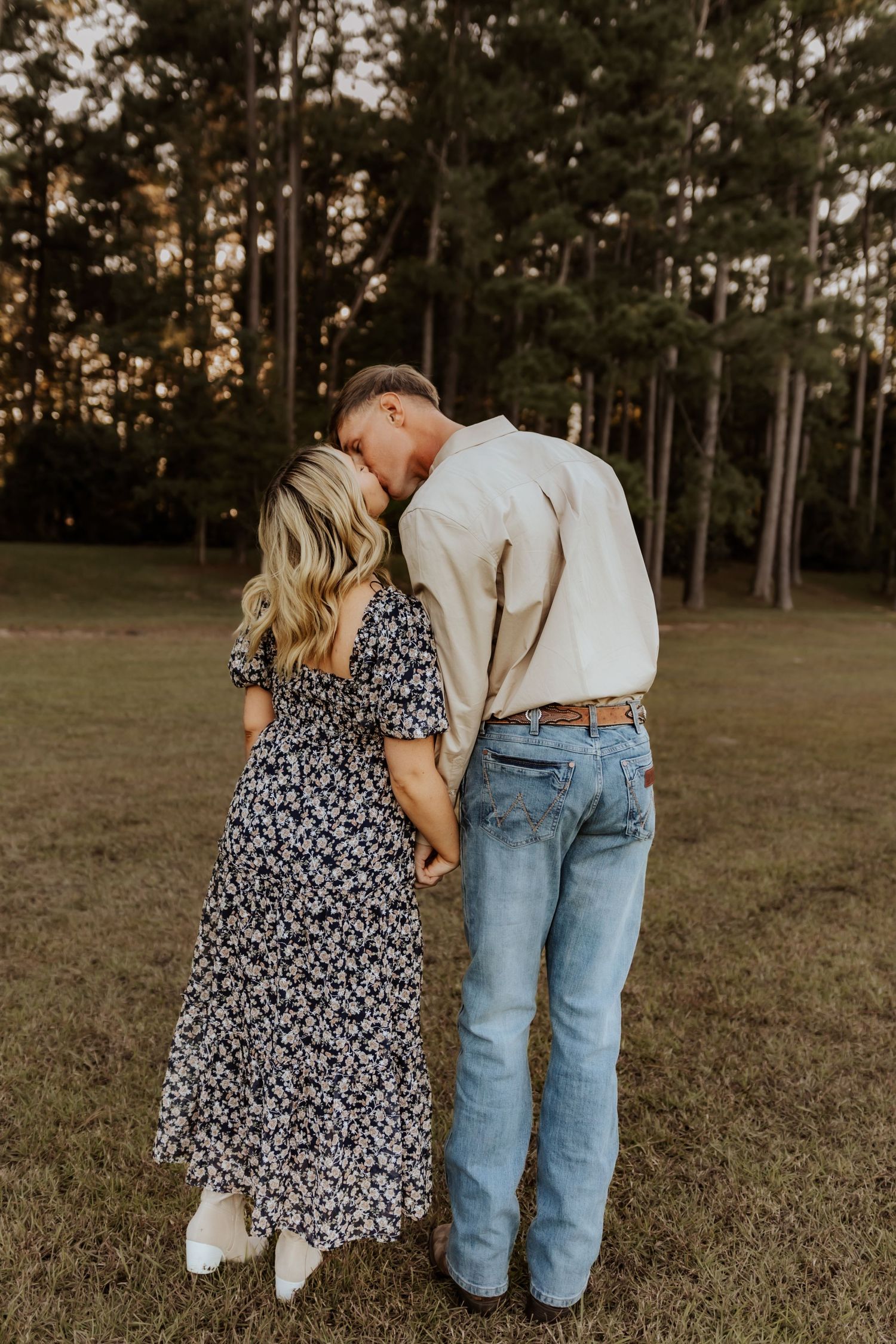 A romantic couple embracing in a grassy field with pine trees at golden hour.