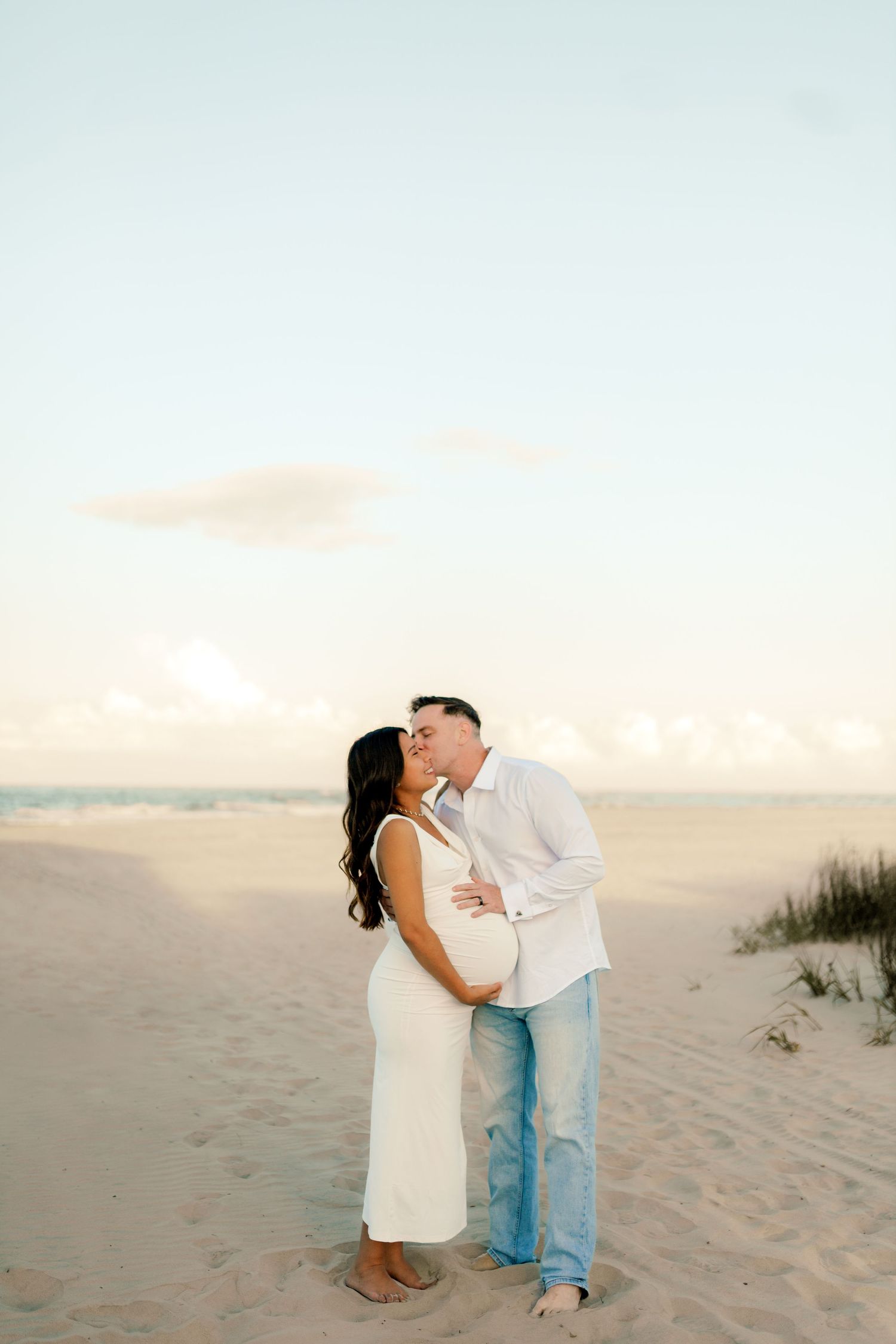 Couple sharing a kiss on sandy beach at sunset, woman in white maternity dress with baby bump, man in white shirt and jeans.