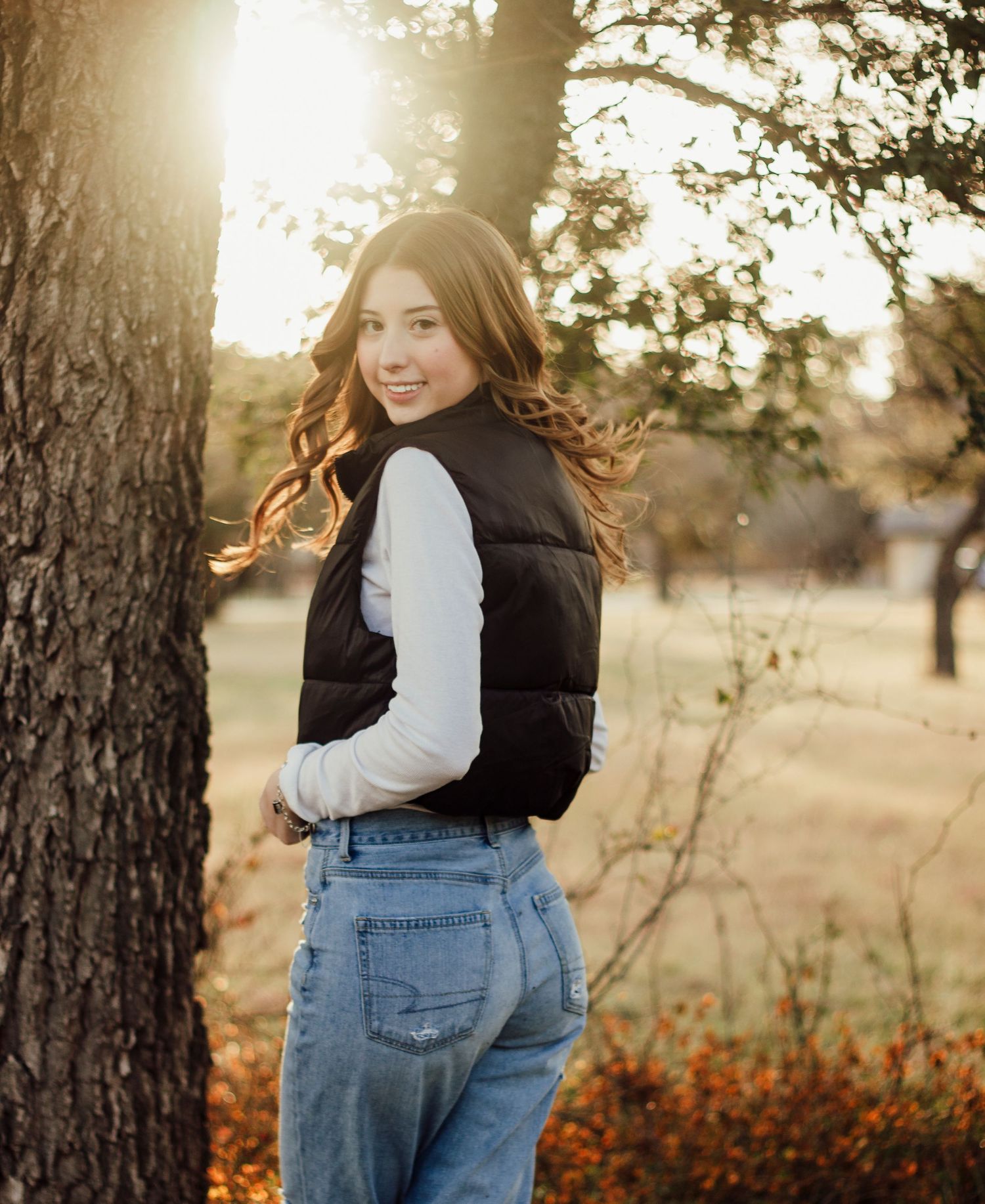 A person in denim overalls stands in a sunlit field with golden light filtering through trees in autumn.