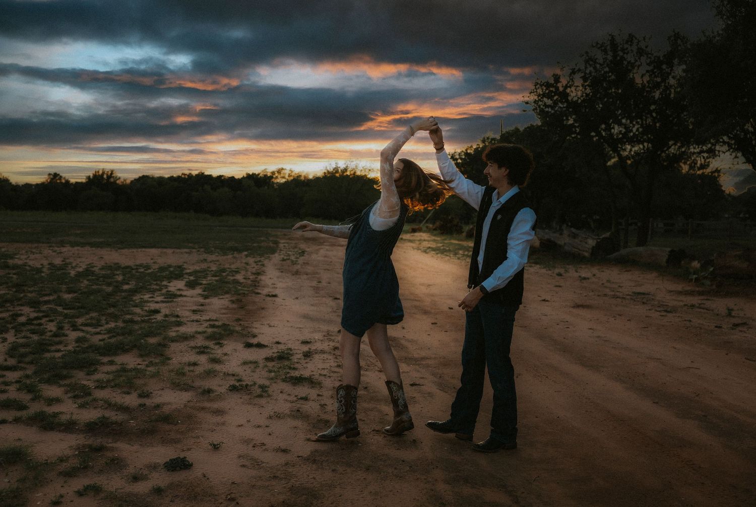 Couple dancing on a dirt road at sunset with dramatic colorful sky and trees in the background.