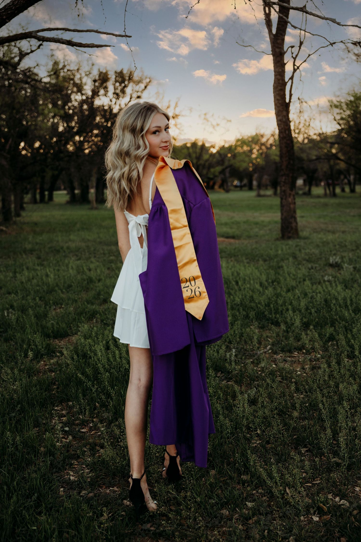 Wylie Senior graduate holds up a purple graduation gown against a sunset-lit field with trees in the background.