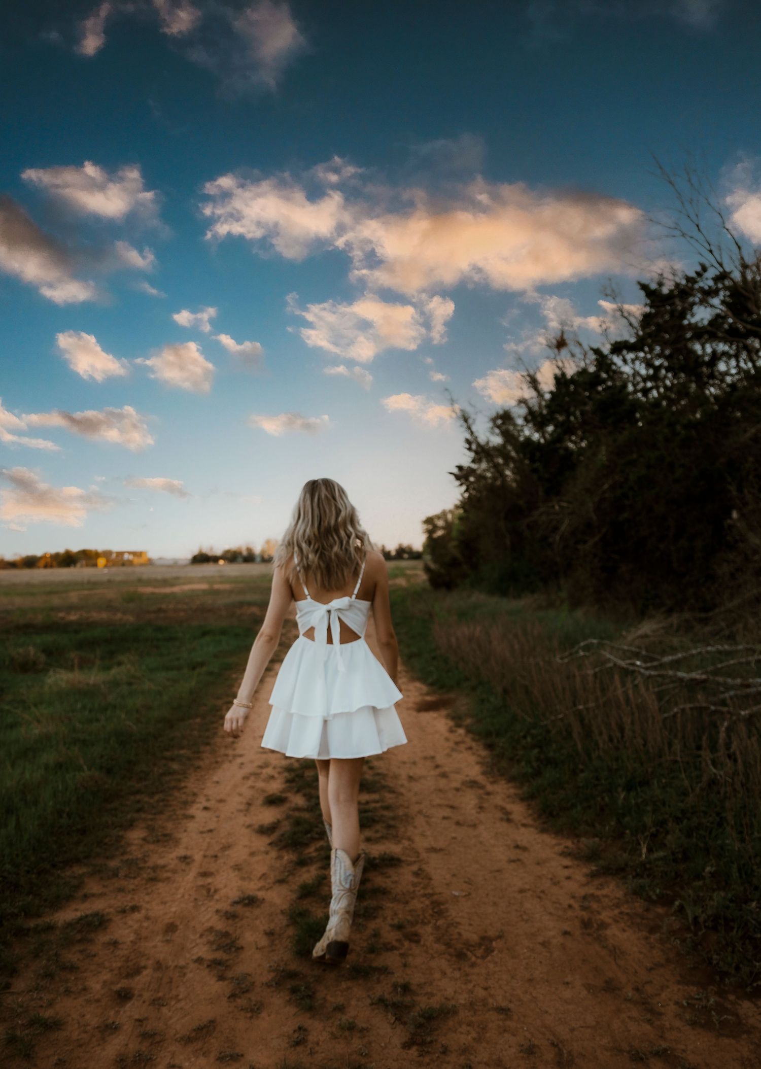 Wylie Senior girl walking barefoot down a dirt path through green fields under a dramatic blue sky. In Abilene Texas