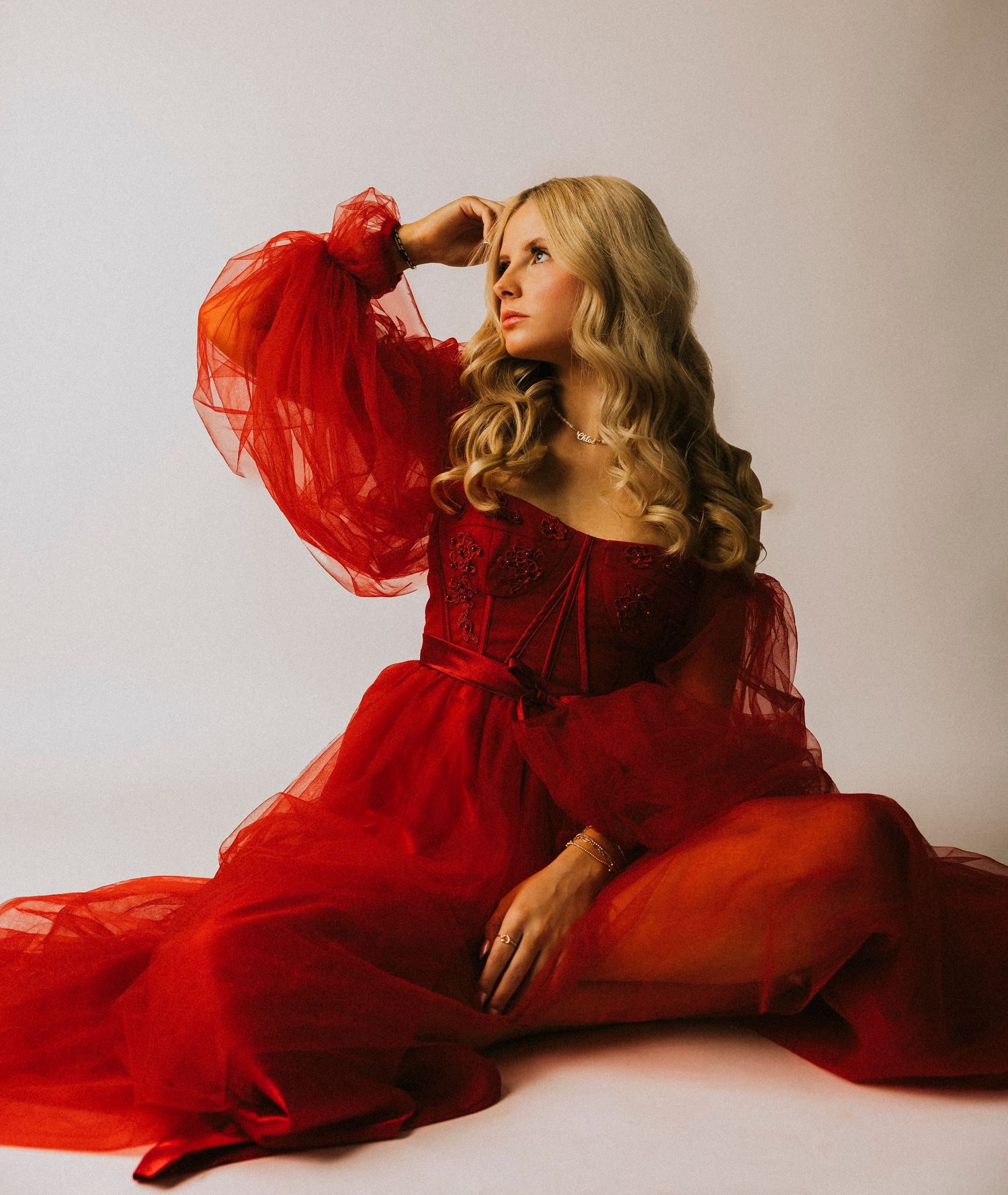 Abilene TX Senior High School girl in elegant red tulle gown with sheer sleeves sits gracefully on floor against white background.
