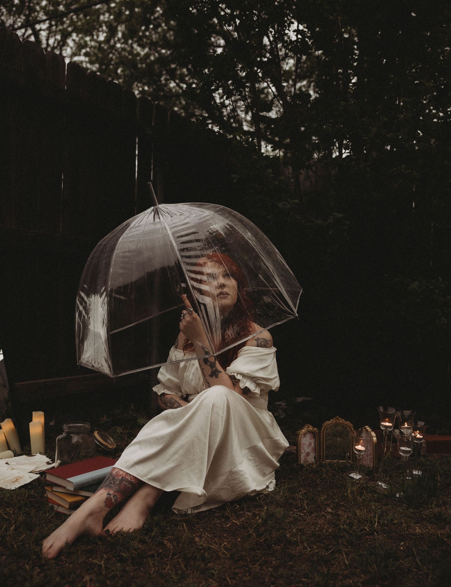 Woman in white dress holding clear umbrella surrounded by candles and jars in a dark, moody outdoor setting.