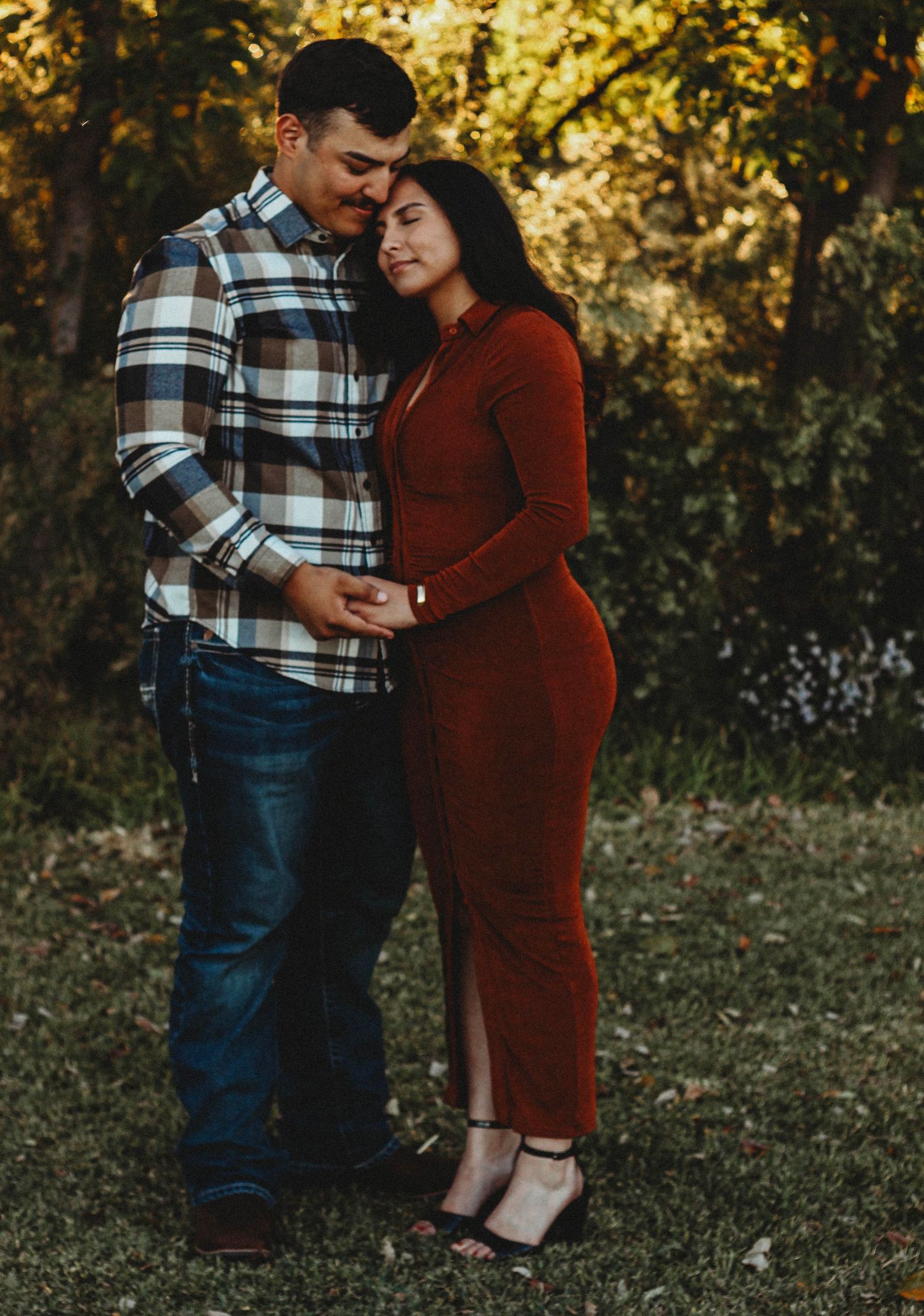 A couple shares a tender embrace outdoors in autumn, man in plaid shirt and jeans, woman in rust velvet dress.