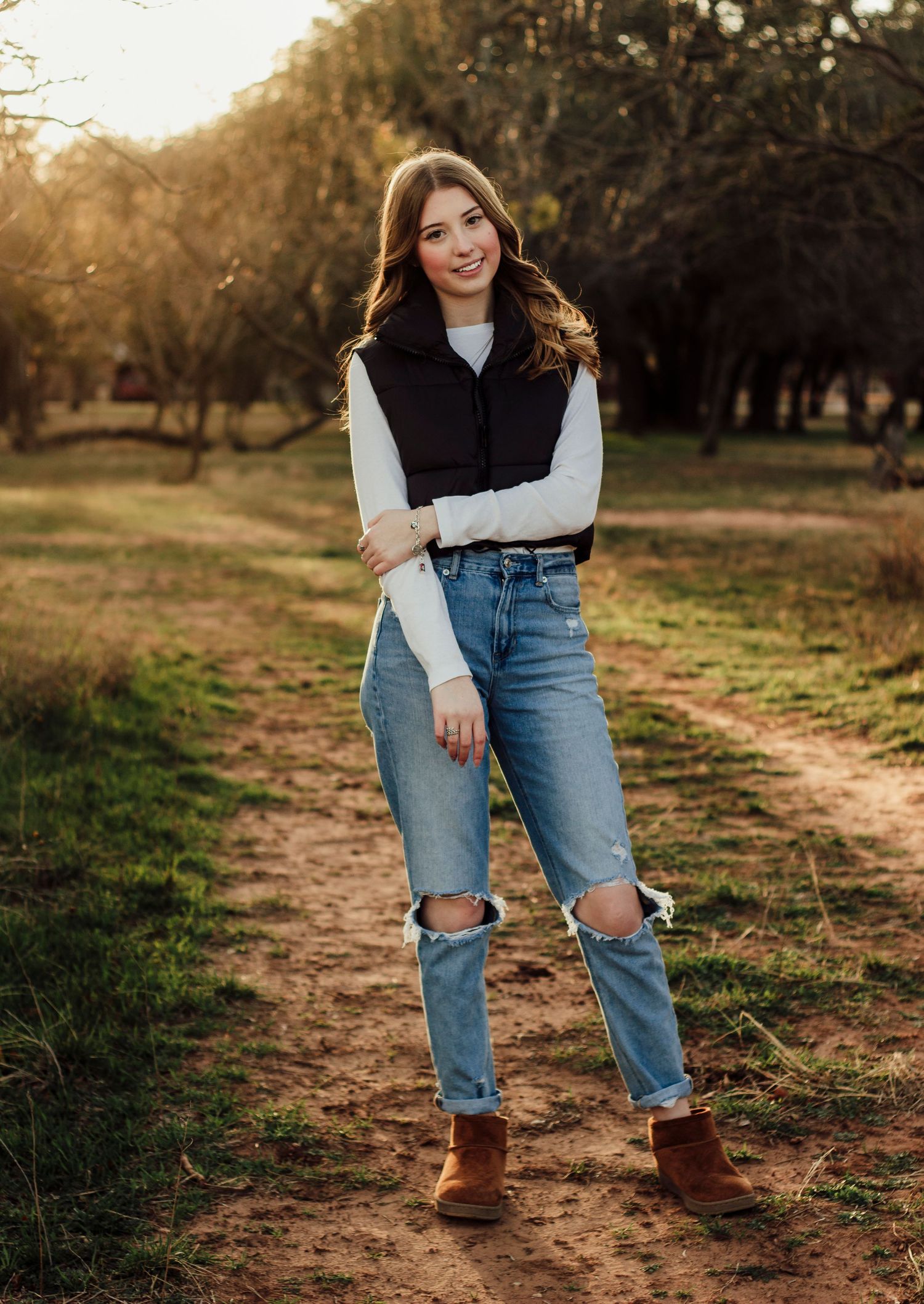 Casual autumn outfit featuring ripped blue jeans, brown suede boots and a black vest on a dirt path at sunset in Abilene TX
