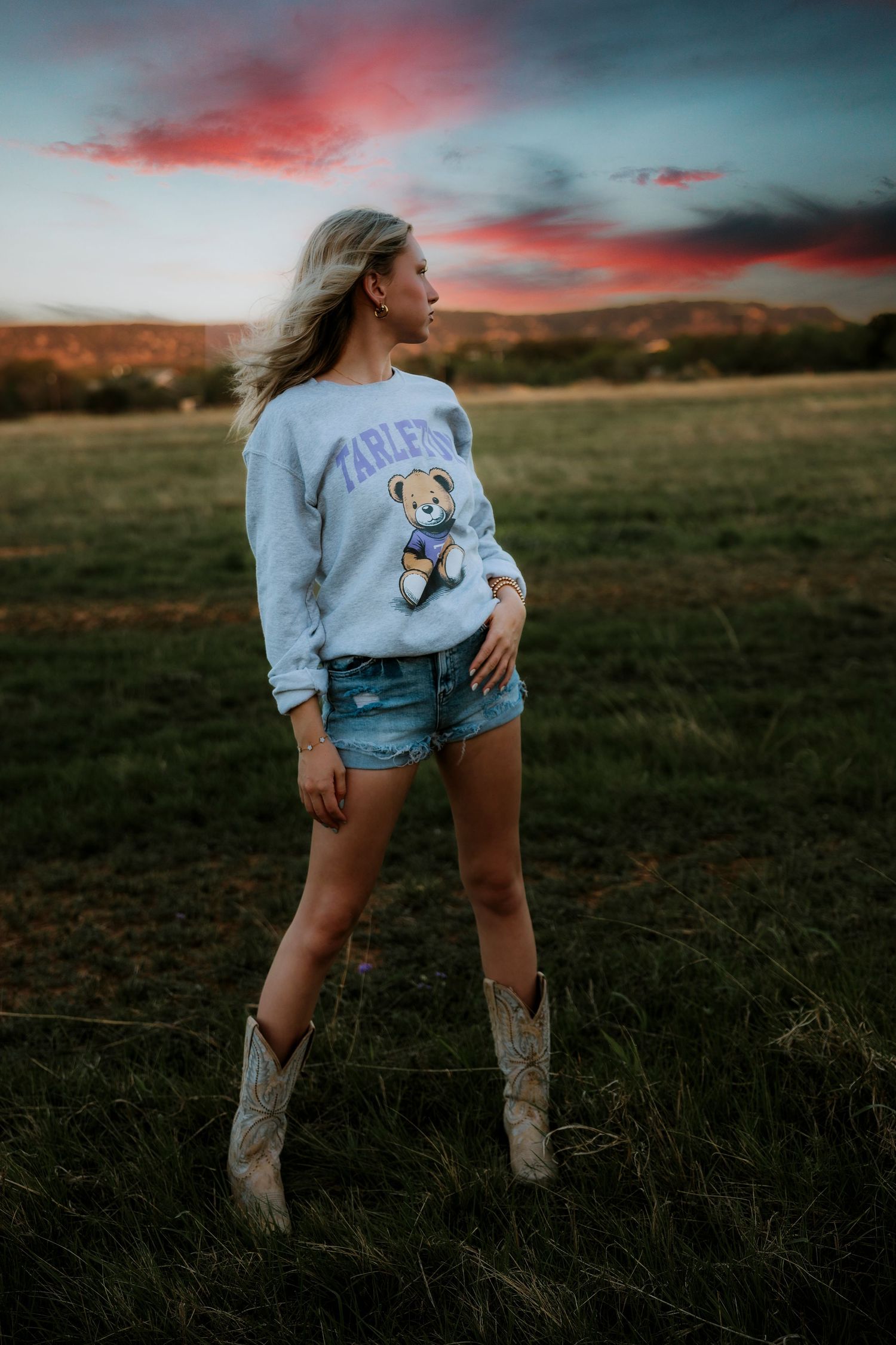 Senior girl  in Tarleton State sweatshirt, denim shorts, and cowboy boots standing in a field at sunset.