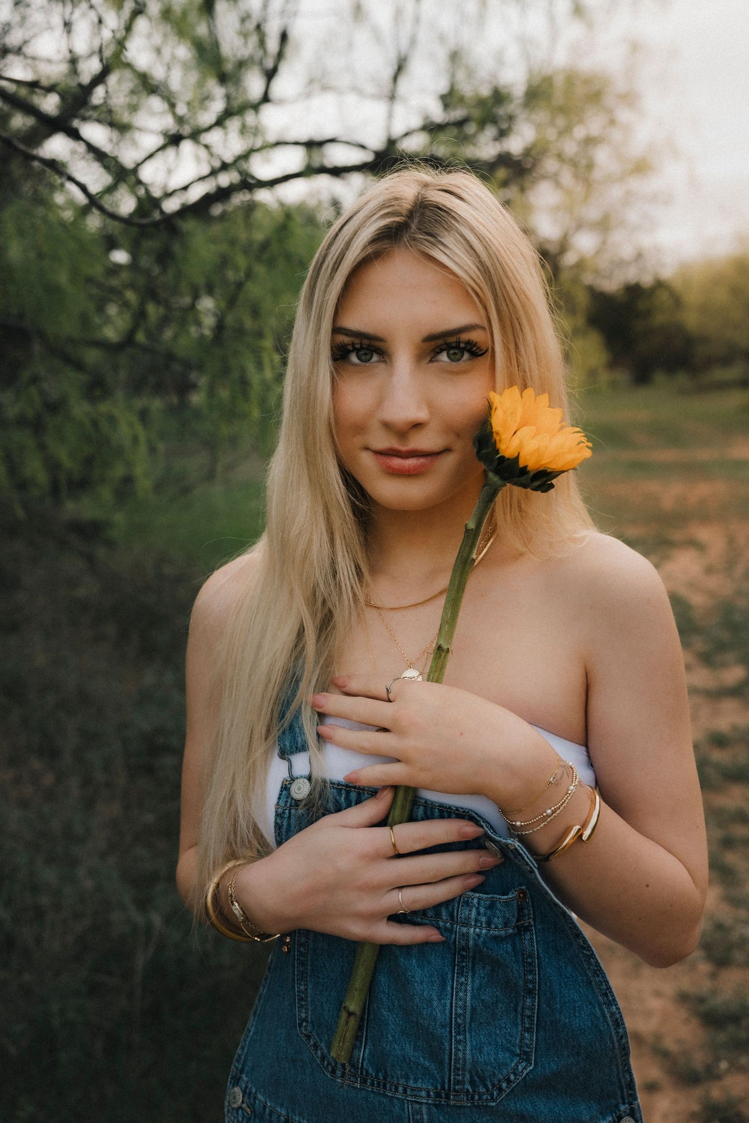 Clyde Senior girl photoshoot denim overalls holding a yellow sunflower outdoors. 90s style edit