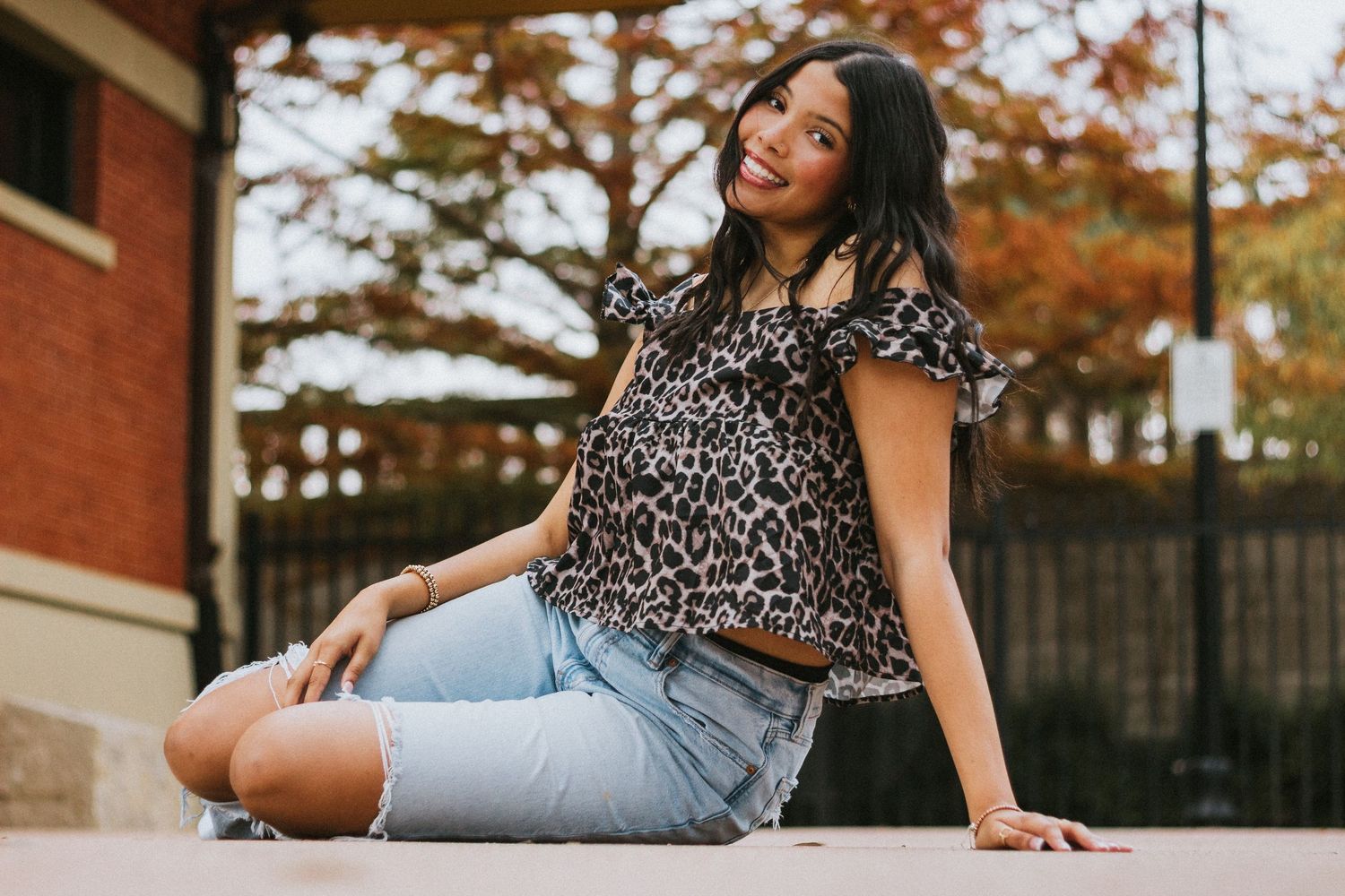 High School Senior in leopard print top and denim shorts sitting outdoors, smiling, with autumn trees in background.