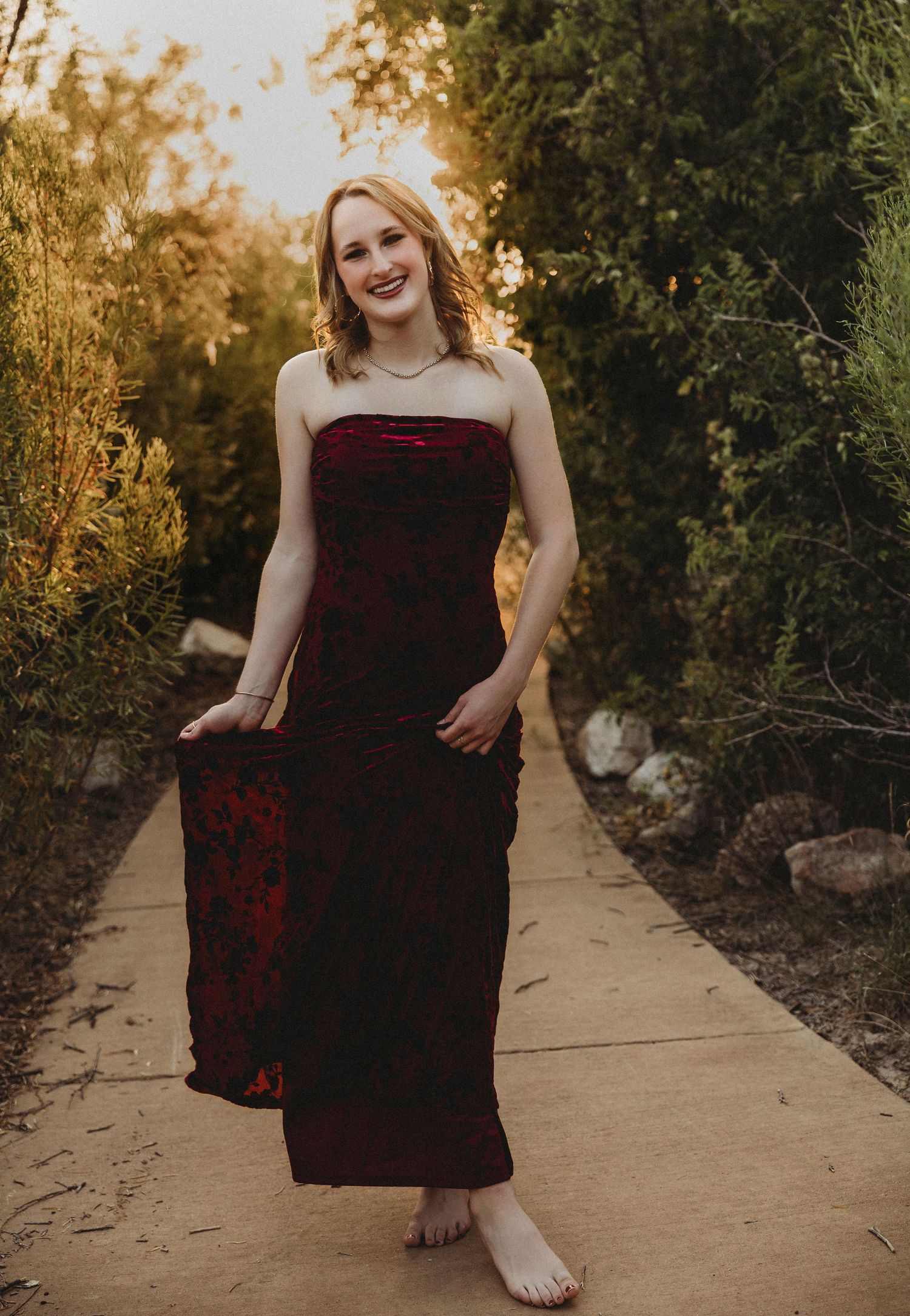 Class of 2027 senior standing on a tree-lined path in a burgundy dress during a natural light senior photography session in Abilene, Texas.