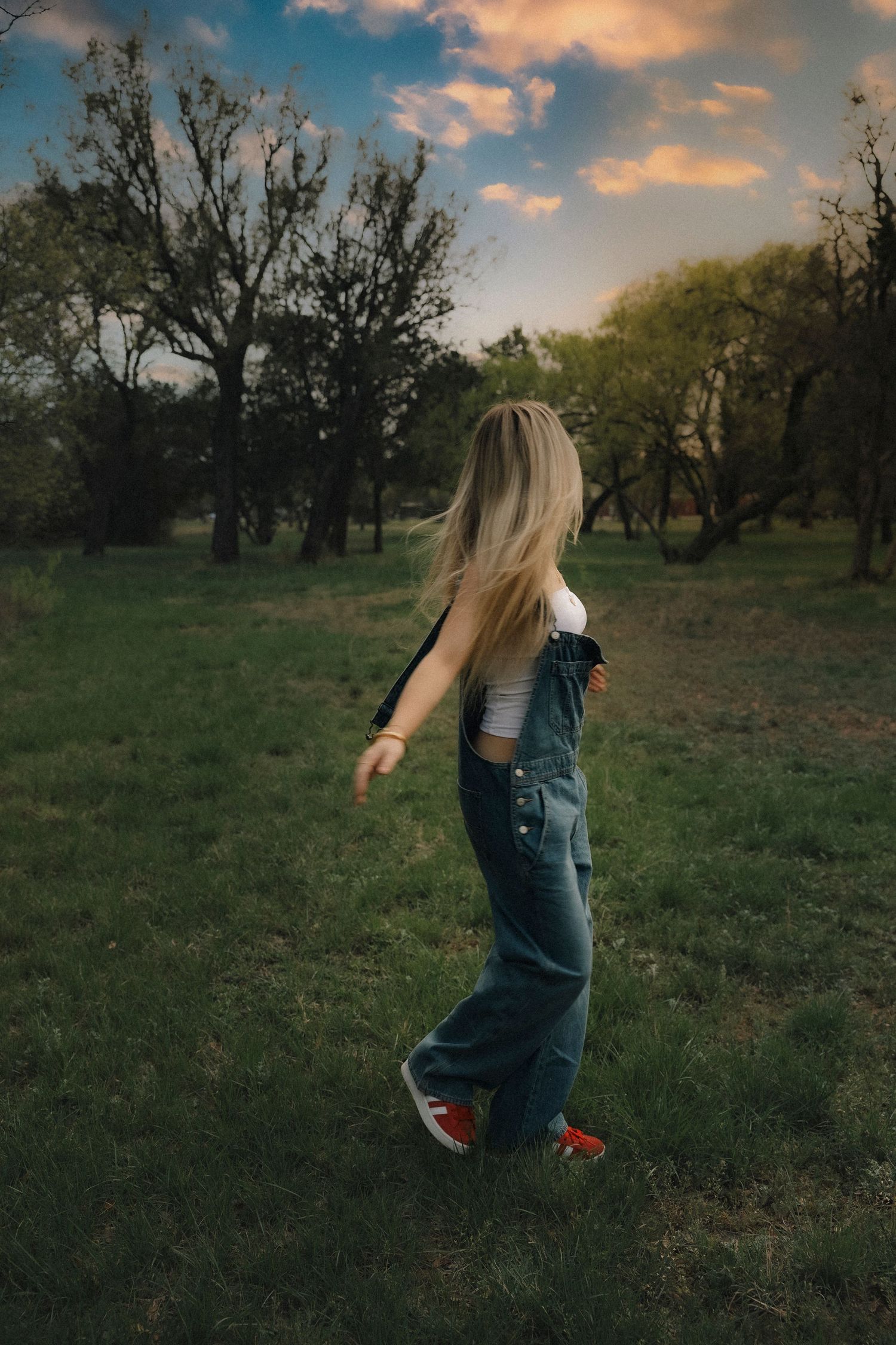 Cylde senior girl  in denim overalls and red sneakers walks through a lush green field at sunset.