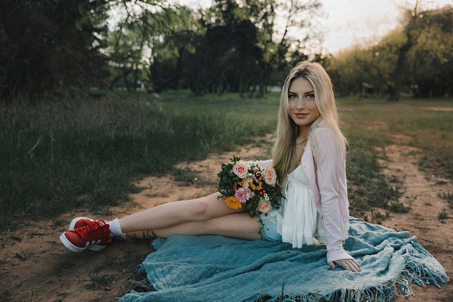 Clyde Texas High School Senior sitting on teal blanket in park holding colorful flower bouquet wearing white dress and red sneakers.