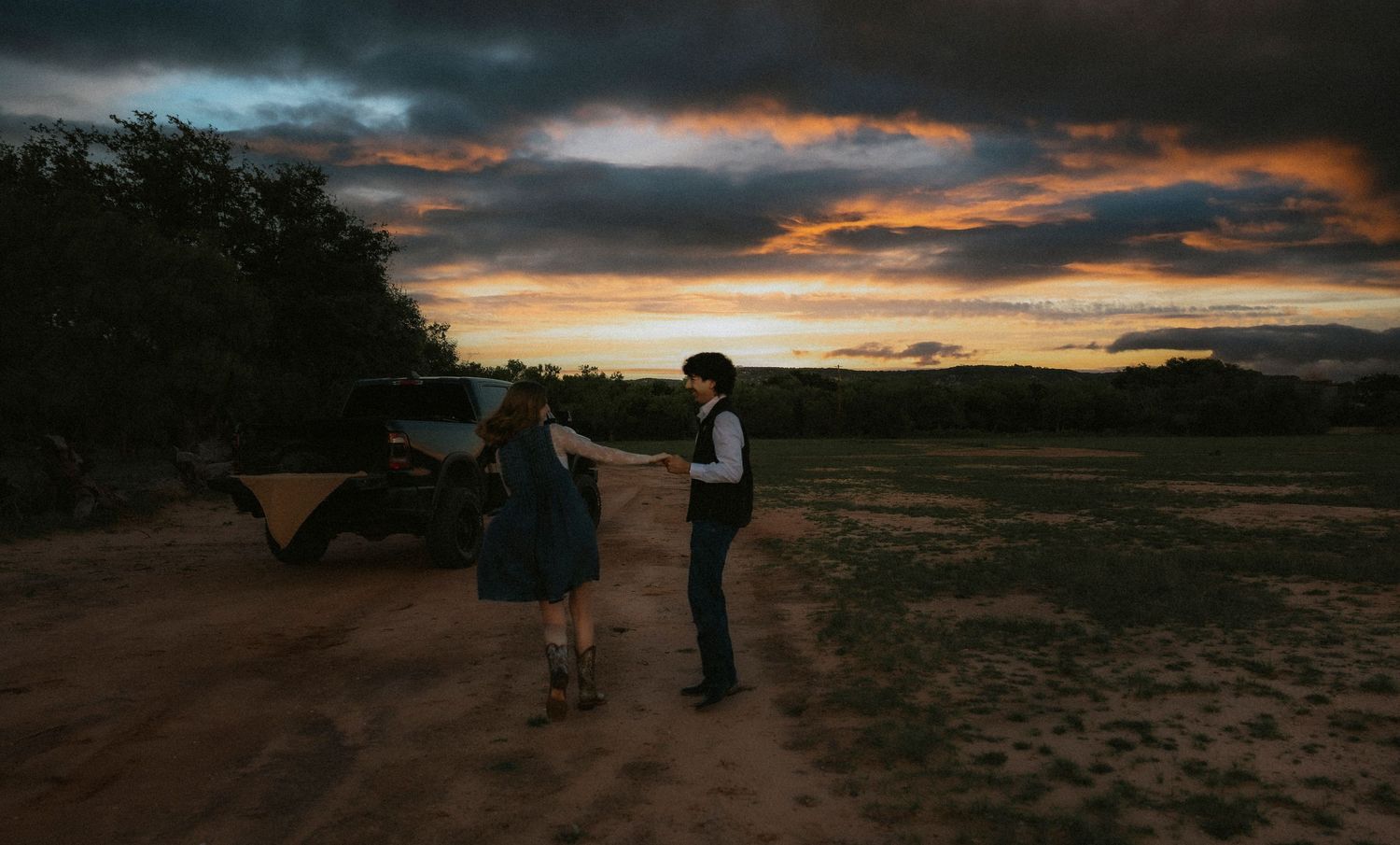 Couple holding hands at sunset in a rural field with dramatic cloudy sky and truck in background.