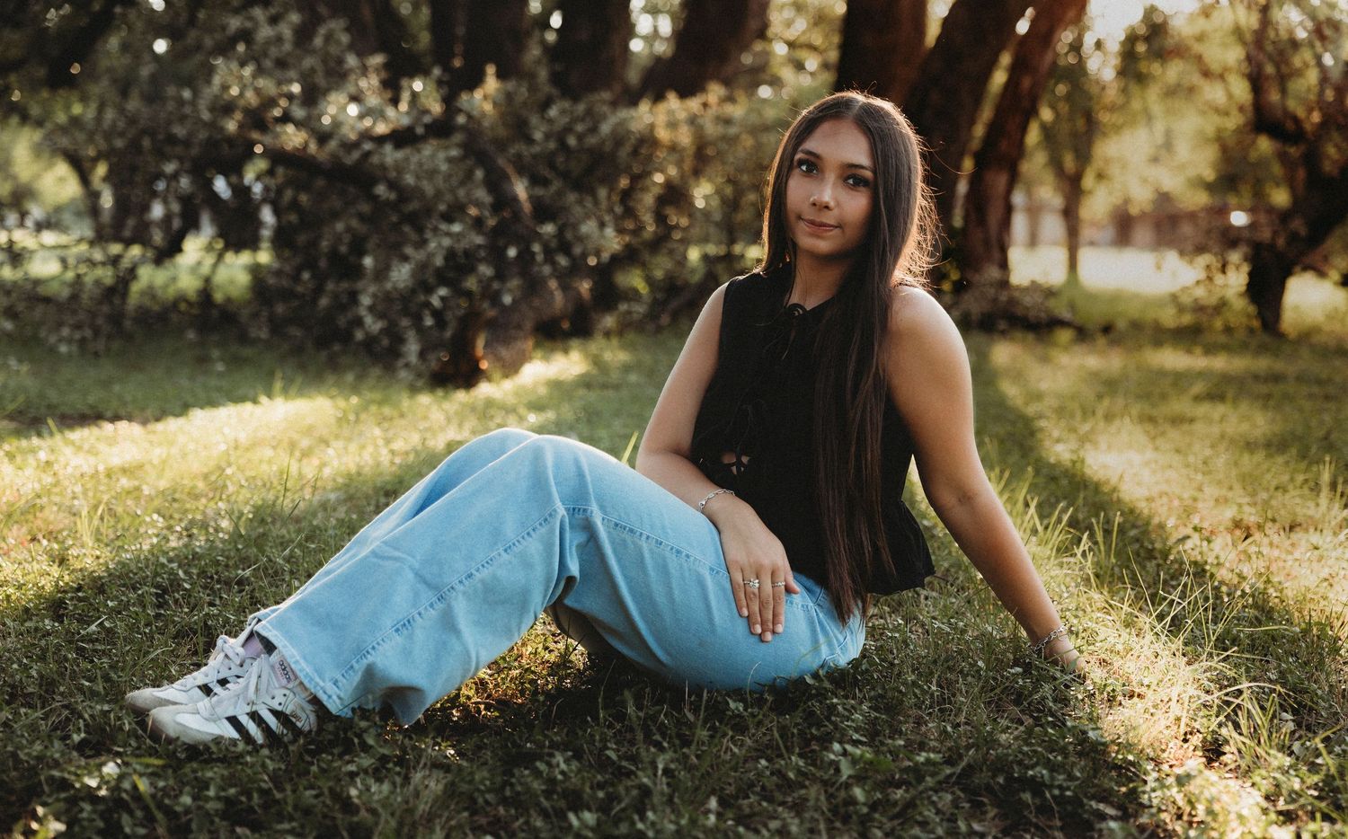 High school senior wearing light blue jeans and a black top sitting in the grass during a golden hour senior portrait session in Abilene, Te