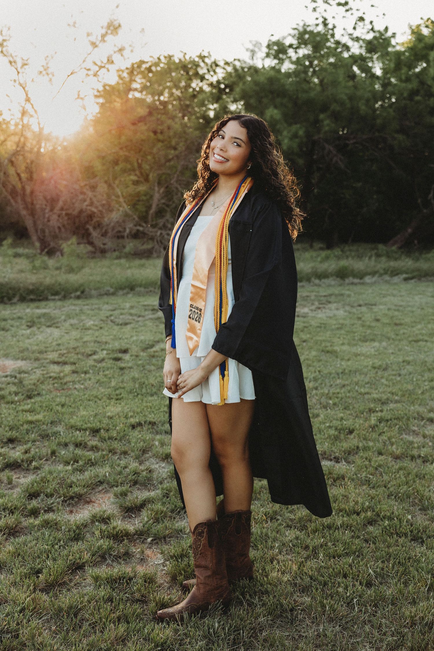 A smiling Abilene High School Senior graduate poses outdoors in a black gown, gold sash, white dress, and cowboy boots at golden hour.